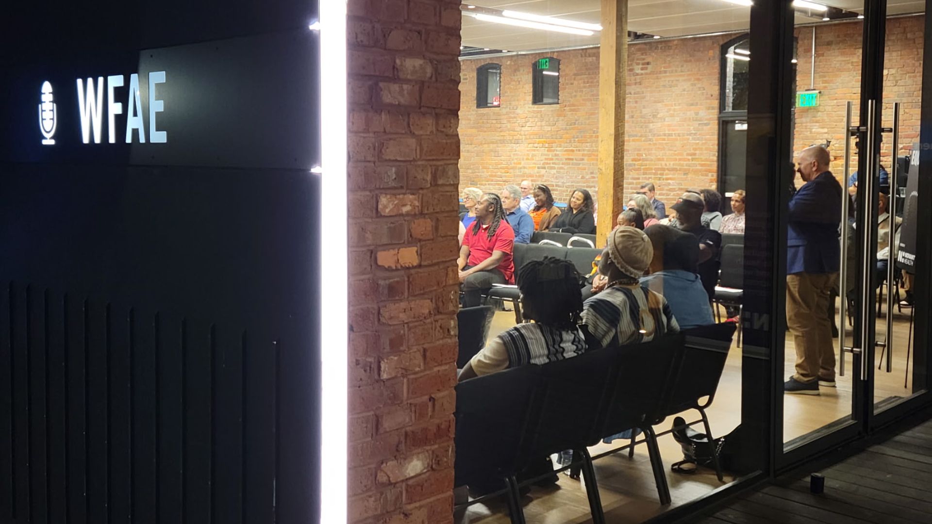 Evening view through glass of a WFAE studio or event space with exposed brick walls; diverse audience seated and listening inside under bright ceiling lights.
