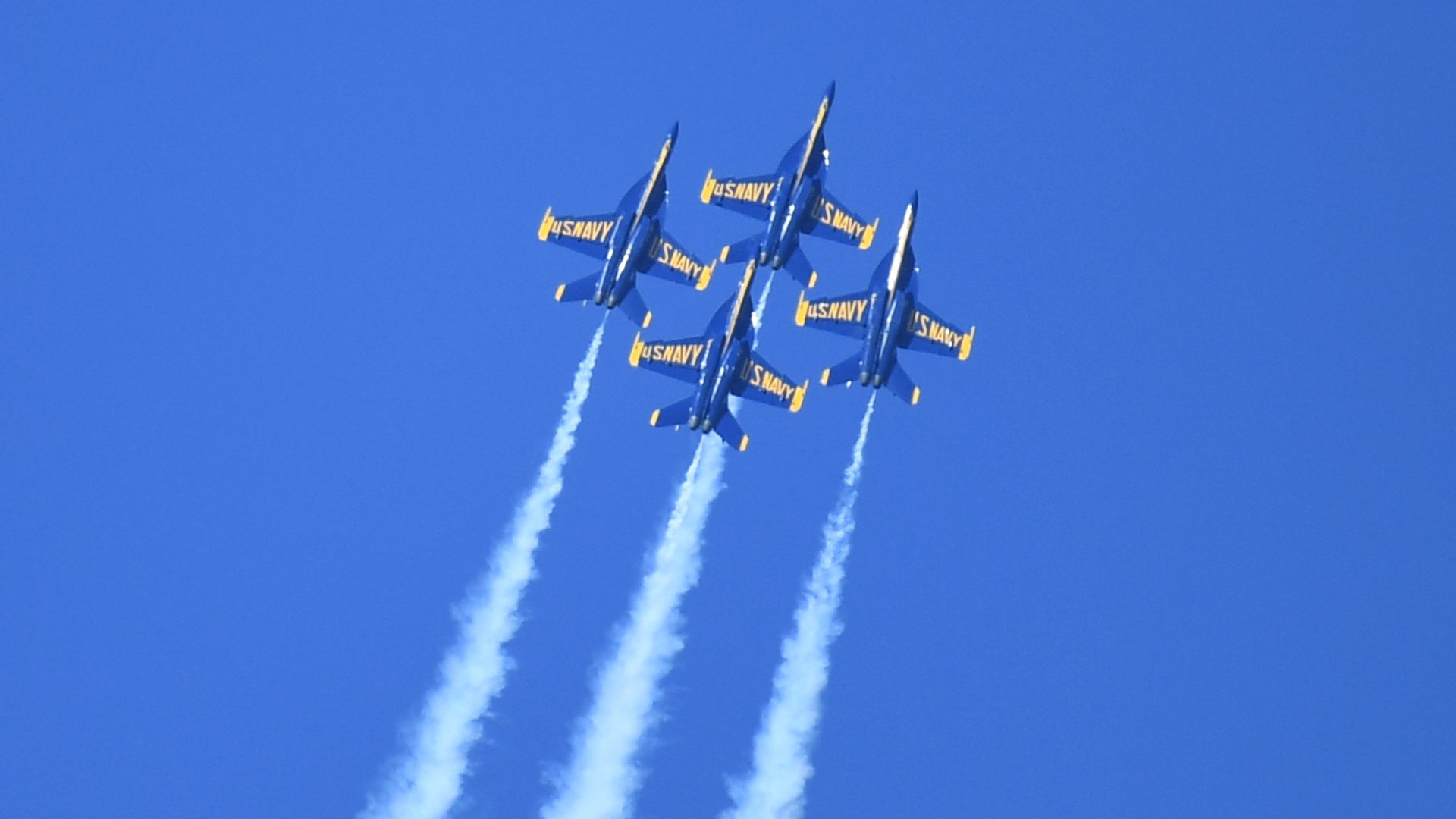 Six US Navy blue and yellow fighter jets fly in a tight diagonal formation, leaving white smoke trails as they streak across a clear blue sky.