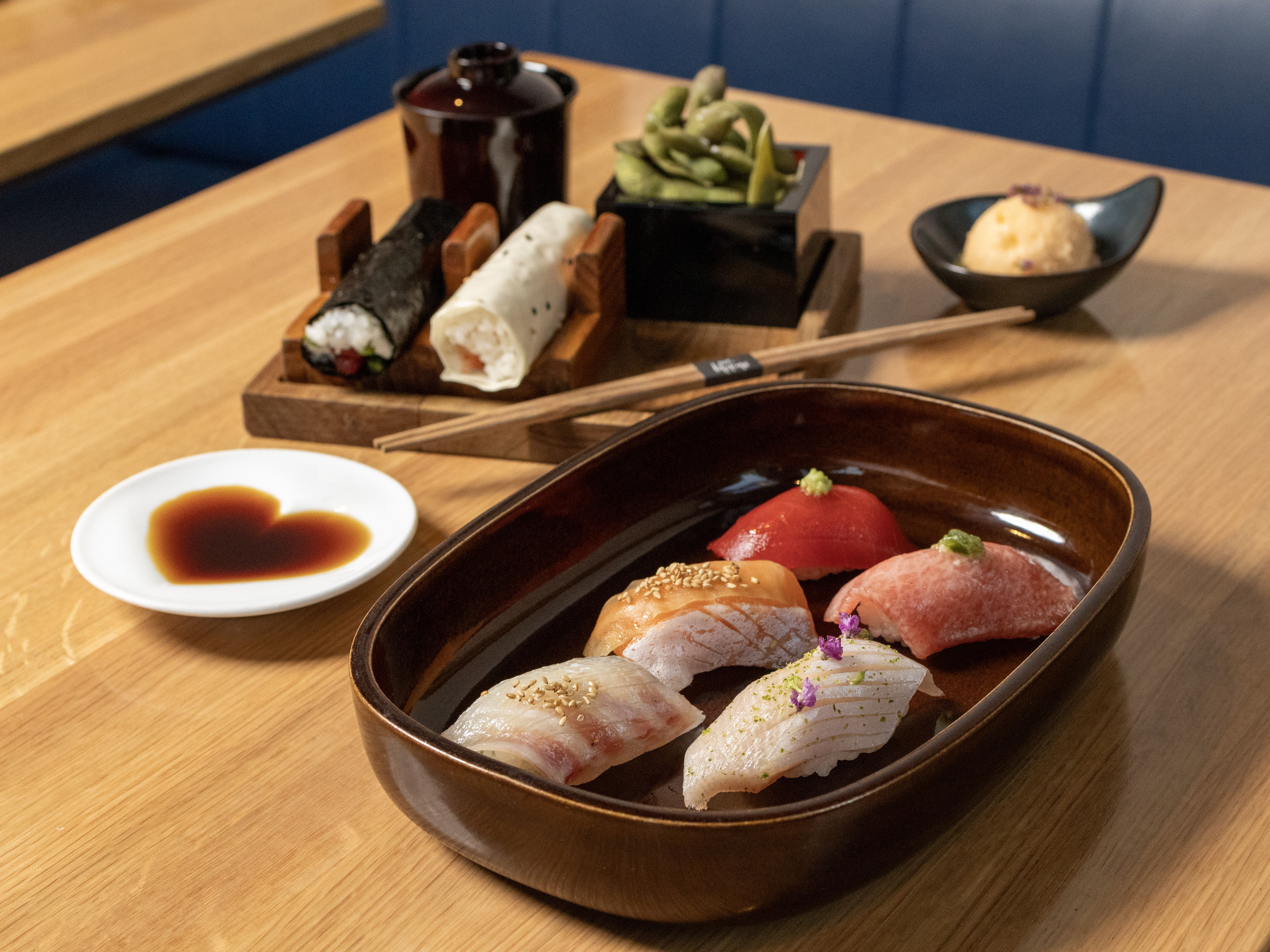 Brown tray with five pieces of sushi garnished with sesame seeds and herbs, wooden table with soy sauce in heart shape, sushi rolls, edamame, and a scoop of orange rice salad.
