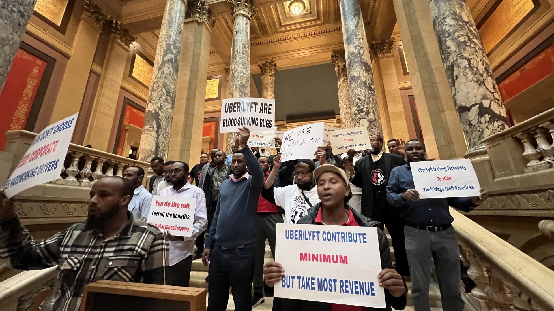 A group of men holding signs saying things like "Uber/Lyft are bloodsucking bugs" and "Uber/Lyft contribute minimum but take most revenue" in an ornate hall with marble columns.