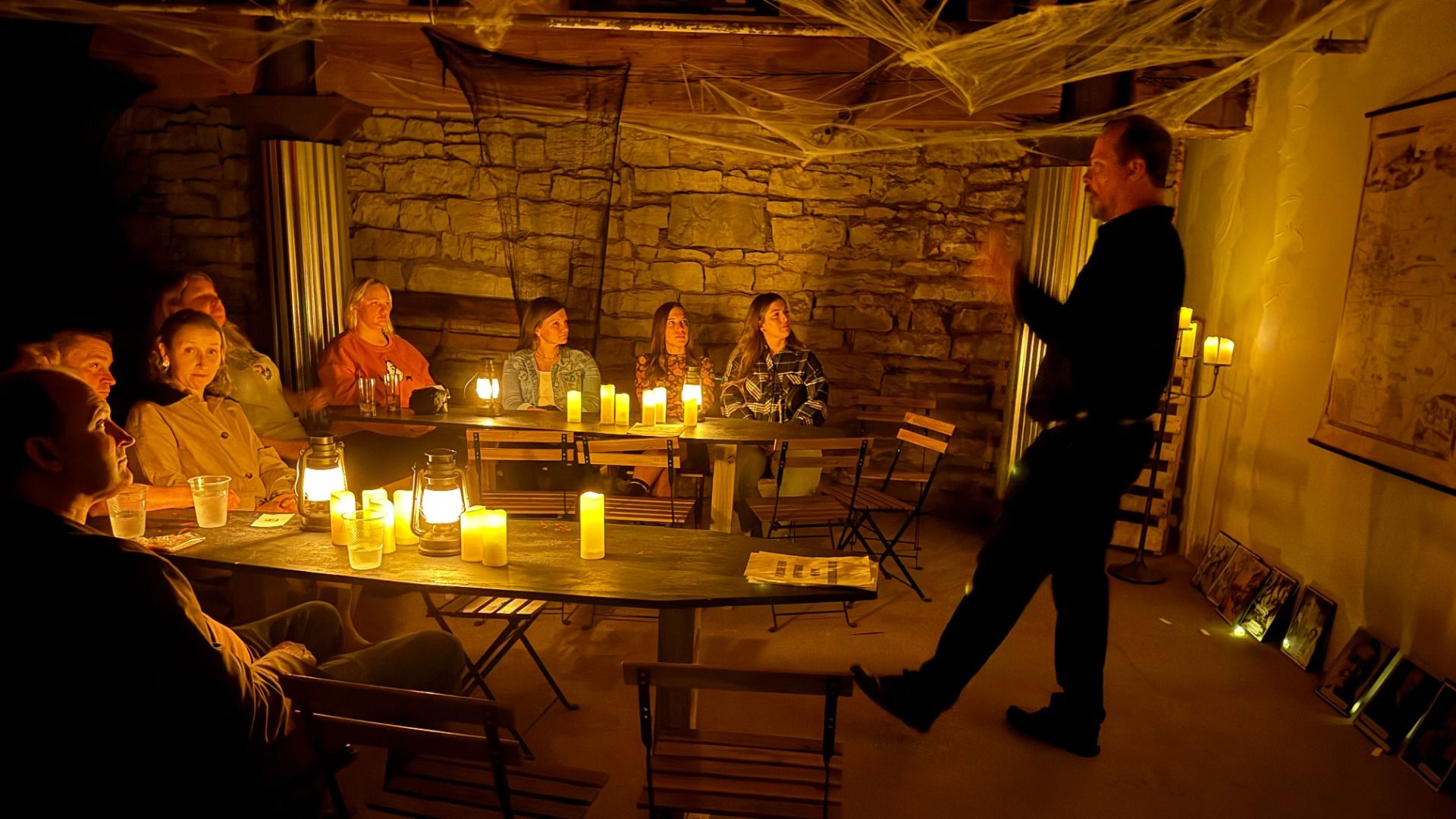 A dimly lit room with stone walls where a man stands speaking to a group of seated people around tables illuminated by candles and lanterns, creating a cozy, intimate atmosphere.