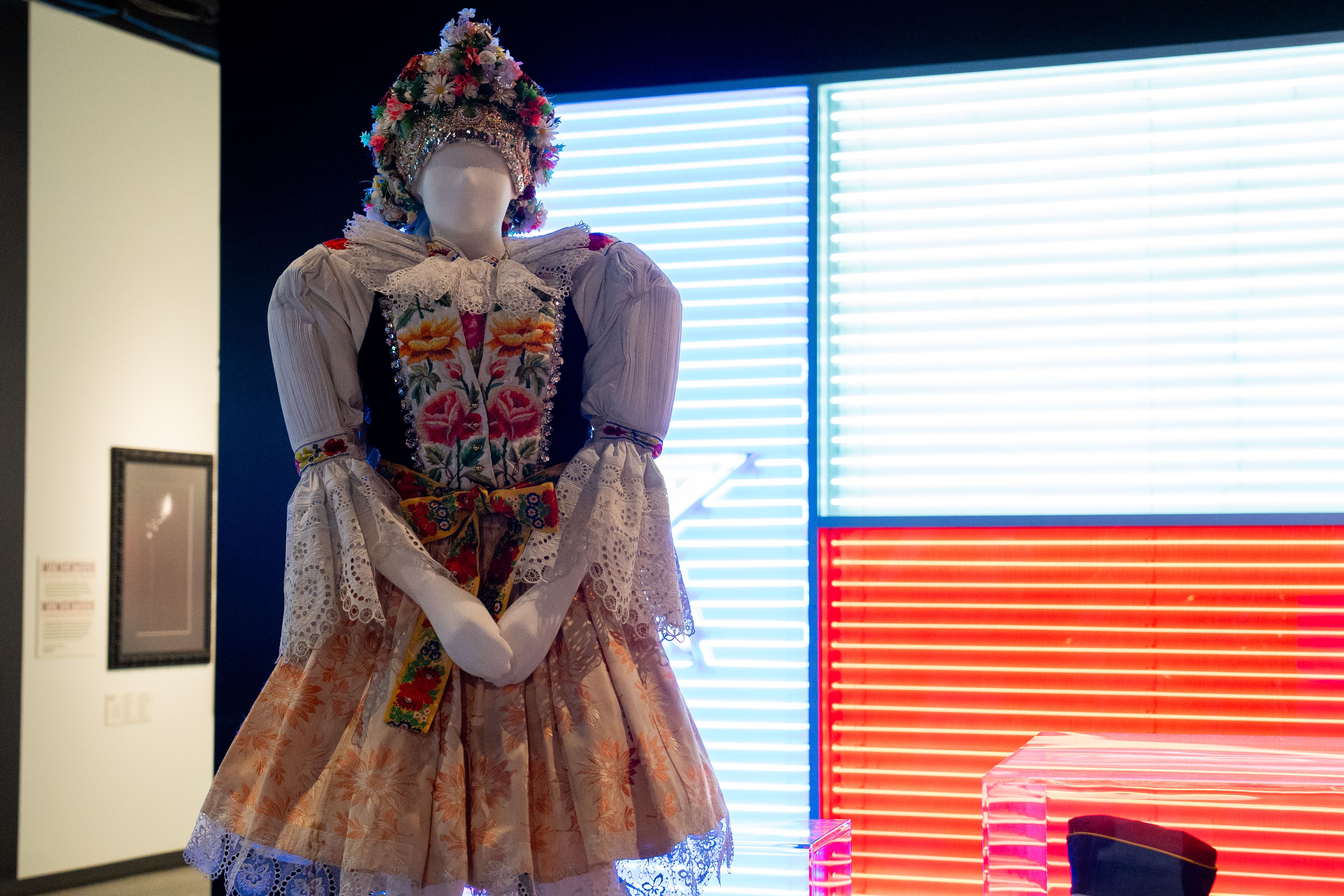 Mannequin in a vibrant traditional folk costume with floral embroidery, lace sleeves, and a flower-adorned headdress, standing in front of bright blue and red neon lights.