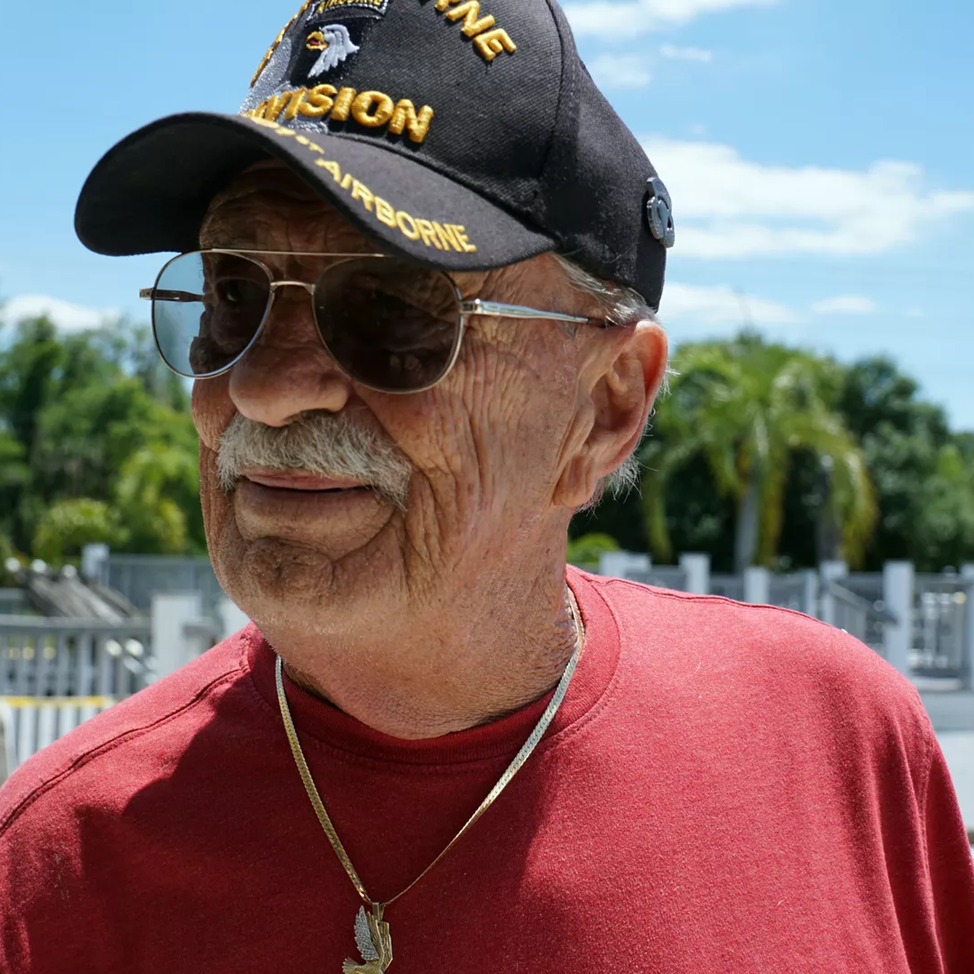 An older man with a mustache and a red t-shirt wearing a hat and glasses
