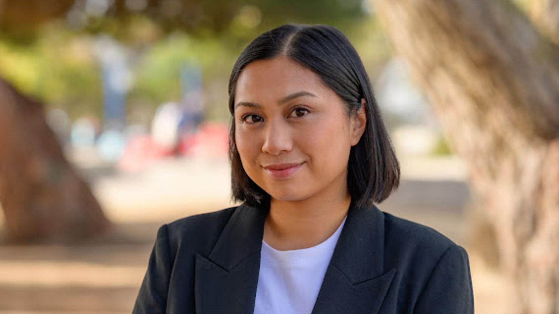 Portrait of a woman with short black hair wearing a white top and black blazer, smiling gently, outdoors with blurred trees and sunlight in the background.