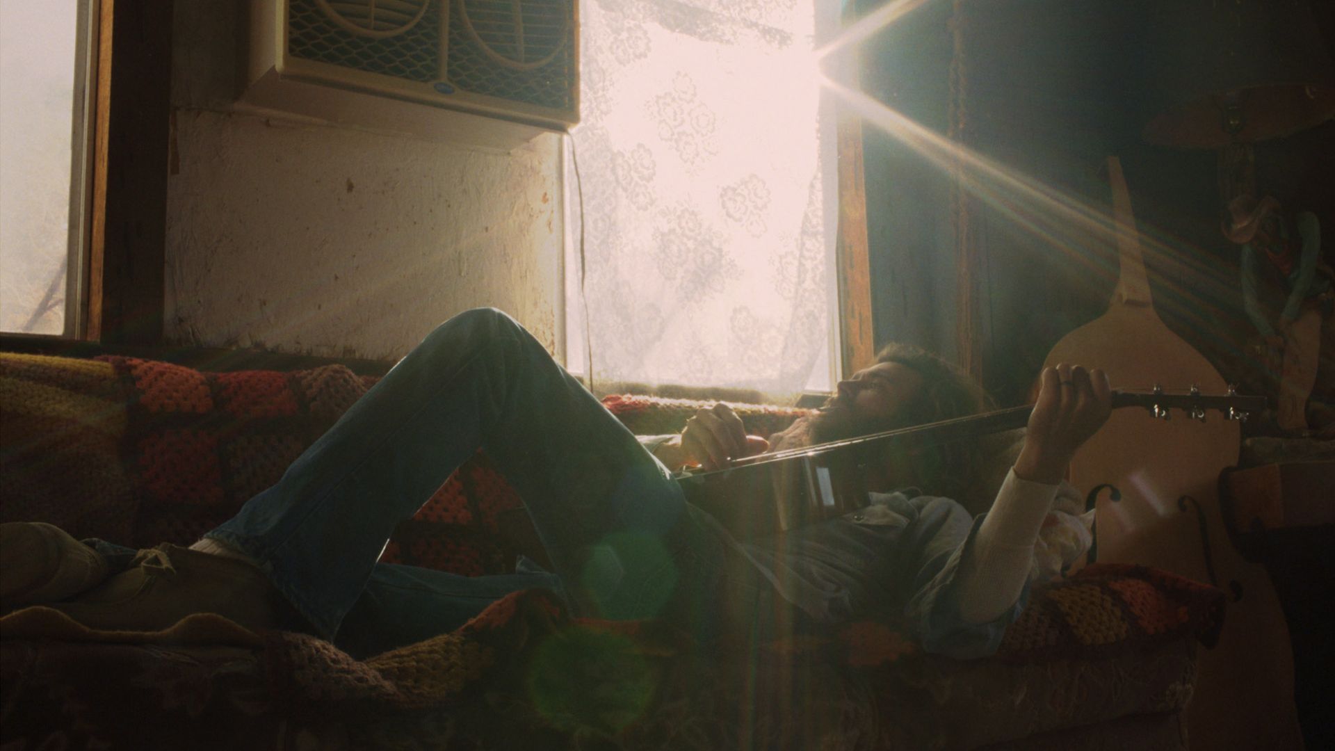 Person lying on a patterned crocheted blanket on a couch, playing an acoustic guitar. Sunlight streams through a lace-curtained window, a dusty room with a guitar leaning upright in the corner.