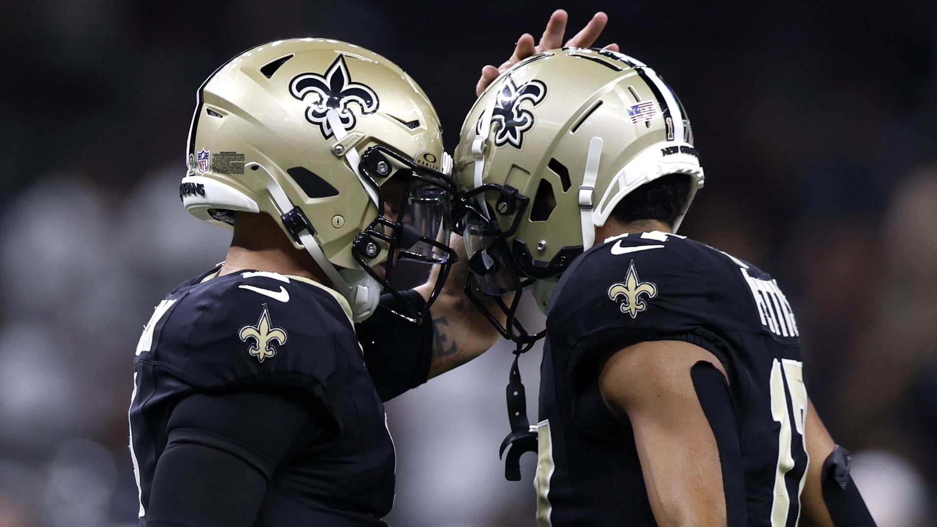 Two New Orleans Saints football players in black jerseys and gold helmets touch helmets during a game, celebrating or strategizing on the field.