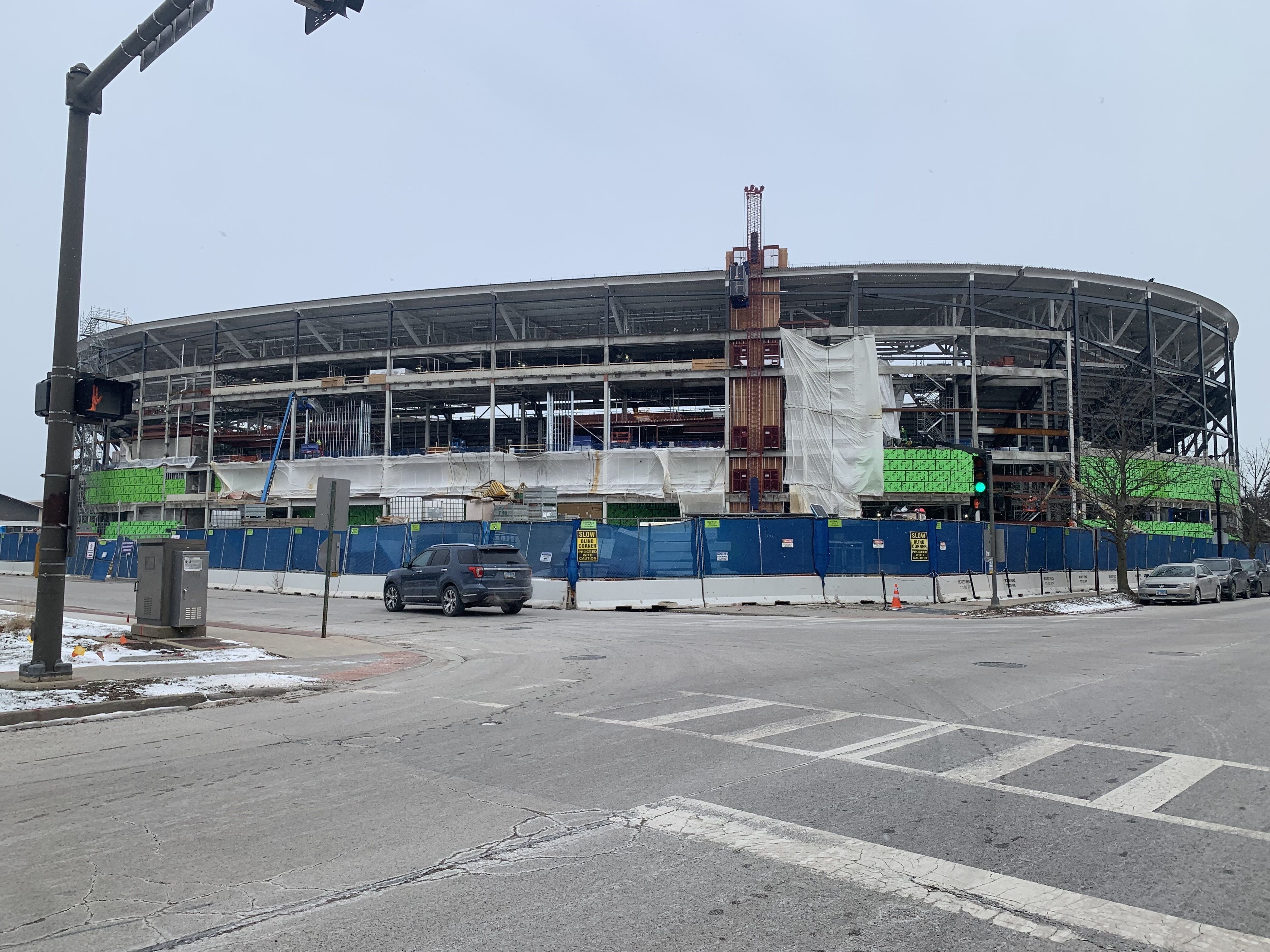 Large circular building under construction with steel framework, white and green construction materials, and blue fences surrounding the site on a gray, snowy day at an urban intersection.