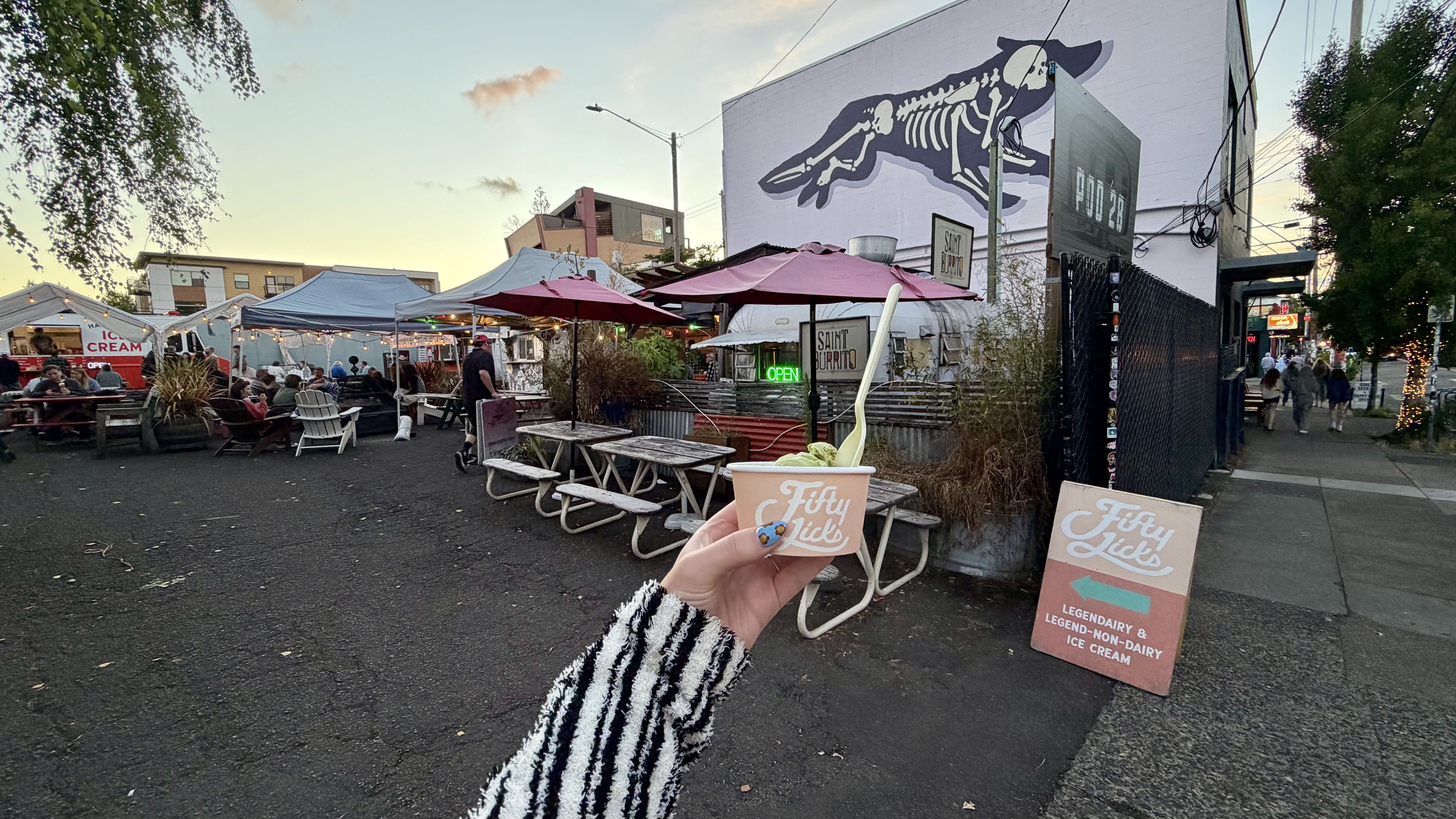 Outdoor market at dusk with string lights and pink umbrellas; people sit at picnic tables under tents, a large skeletal animal mural on a pink wall, and a hand holding a Fifty Licks ice cream cup in the foreground.