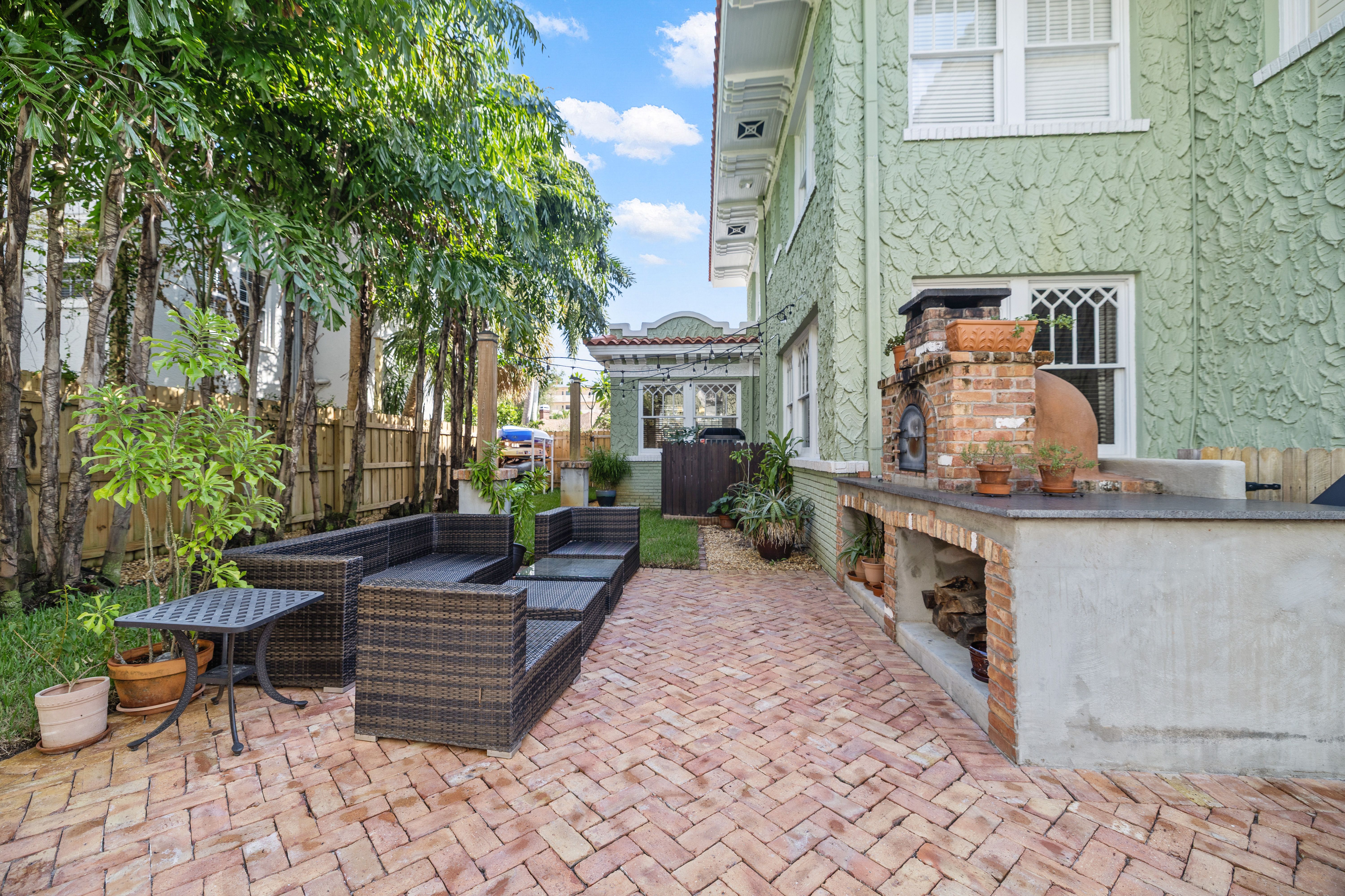 Outdoor patio with brick flooring, black wicker sectional seating, small black table, green potted plants, tall trees, and a brick pizza oven attached to a green textured house under a blue sky.