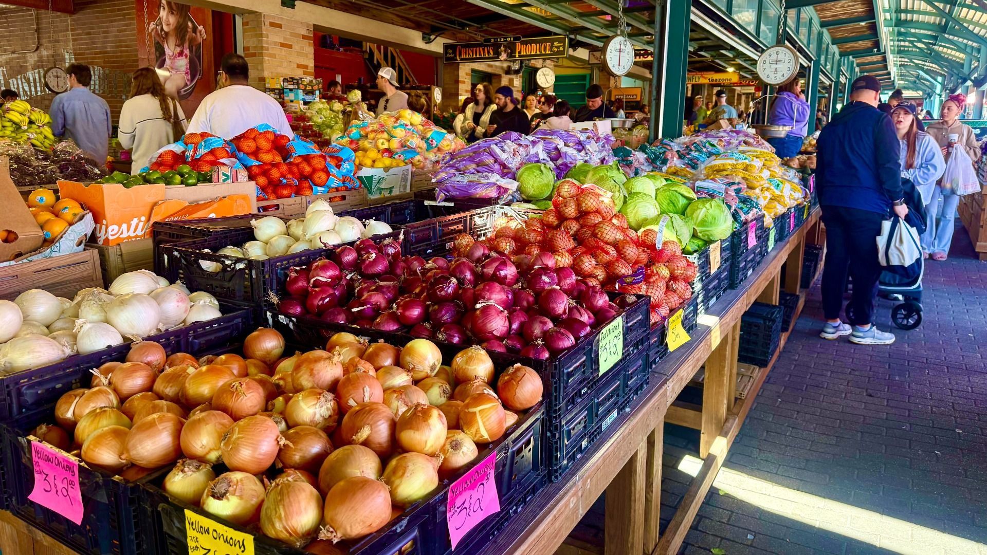 Busy market stall with crates of colorful produce: yellow, white, and red onions; green cabbage; peppers; tomatoes; bags of fruit. Shoppers browse under a brick market canopy.