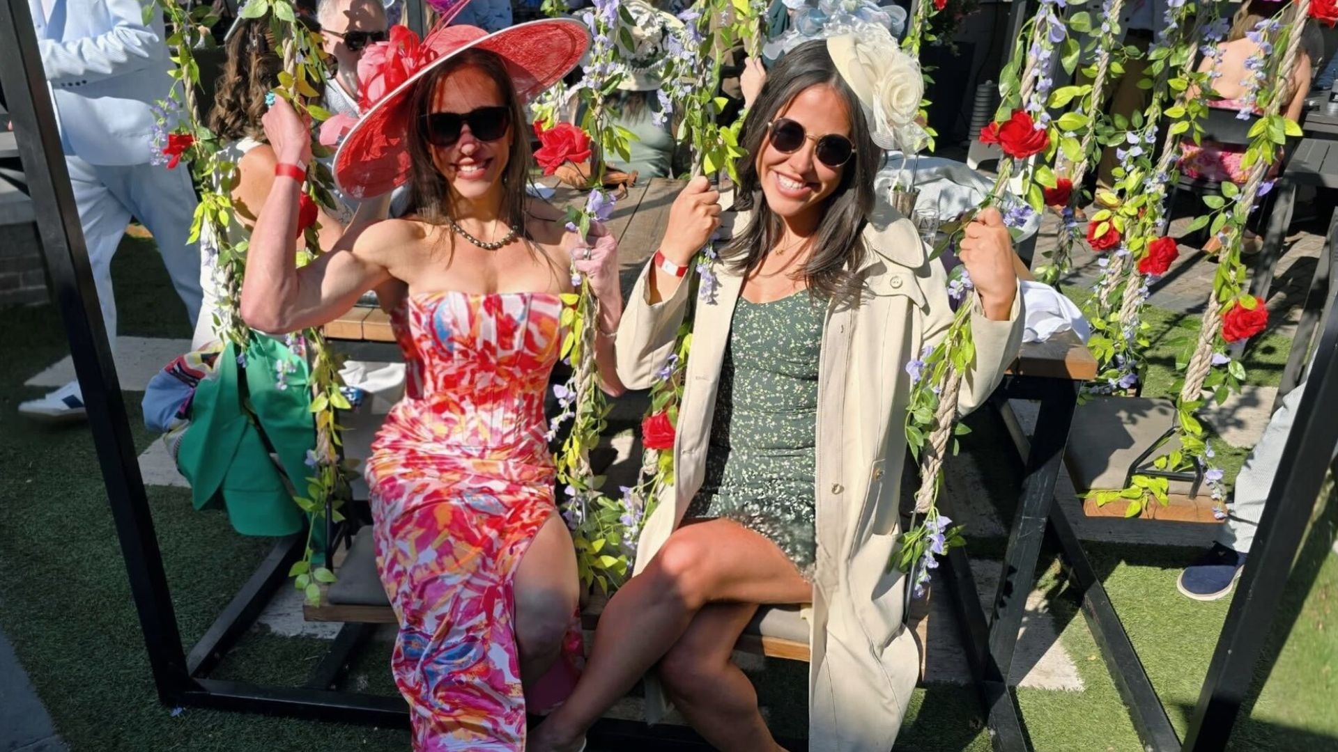 Two smiling women on a flower-decorated swing at an event. One wears a red strapless dress and large hat; the other a green dress with a beige coat and white fascinator, with vines around.