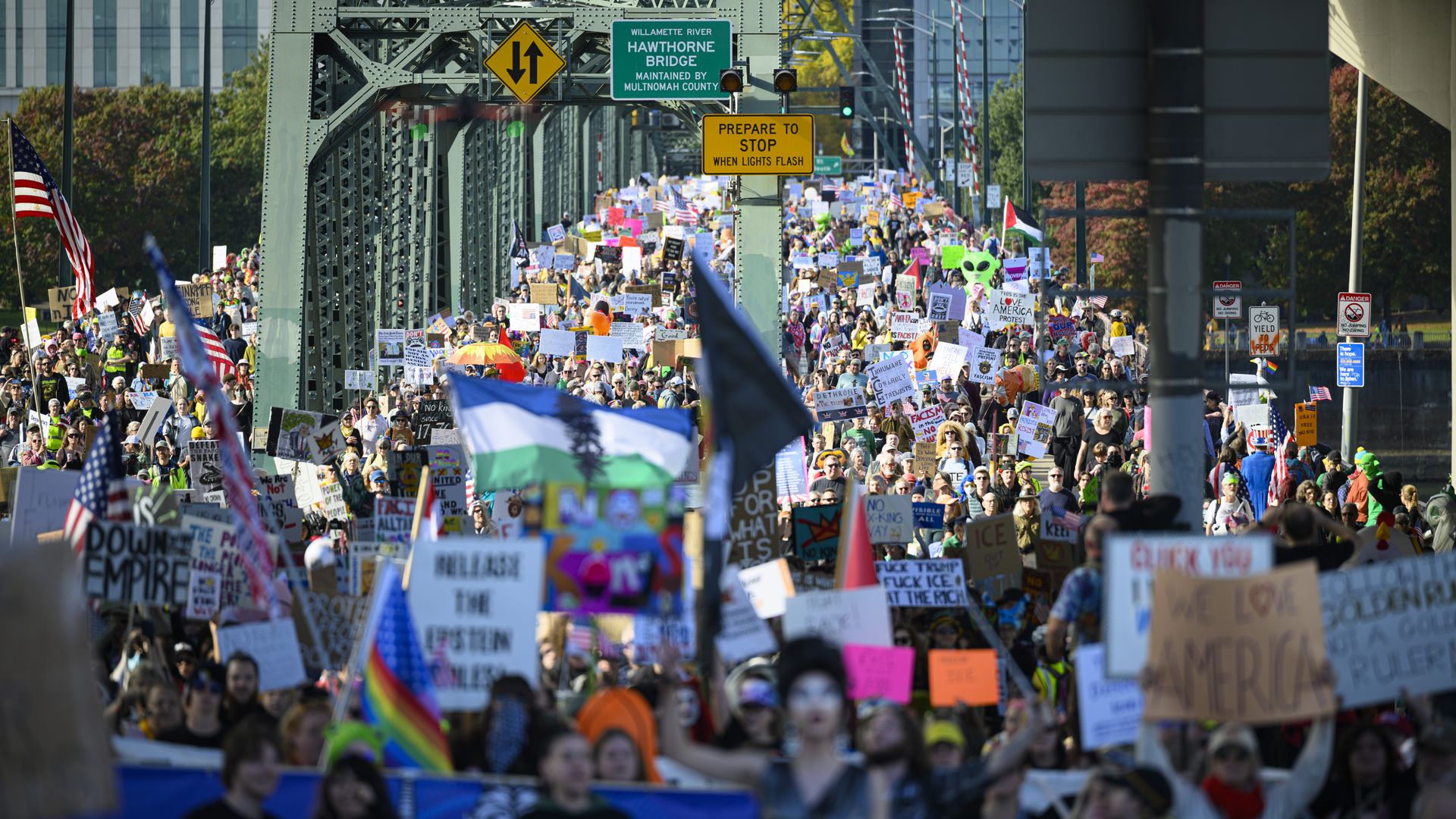 A dense crowd of protesters fills a green metal bridge, waving American flags and holding colorful signs. Overhead signs note Hawthorne Bridge and a yellow warning about stopping.