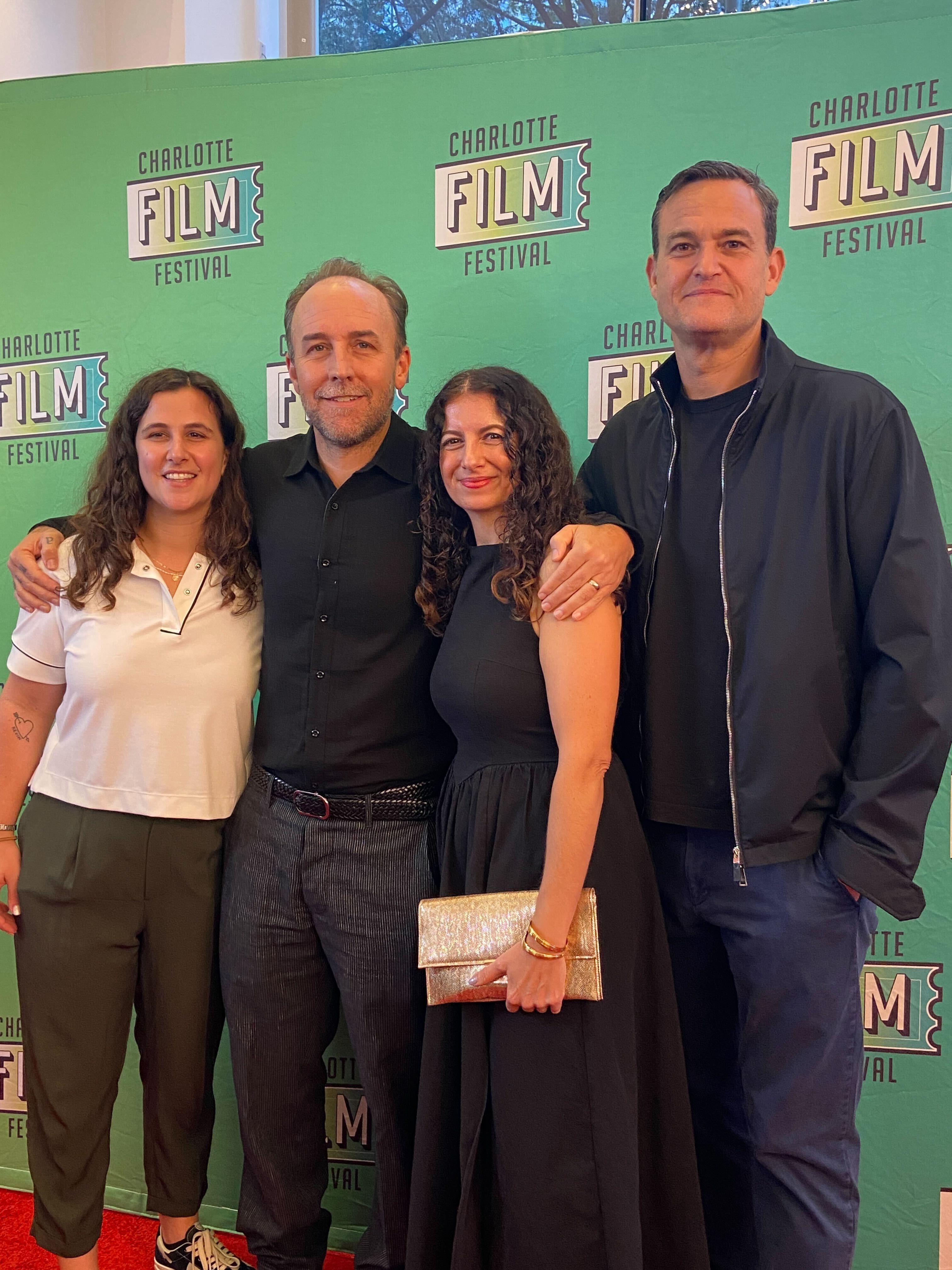 Four people standing close together on a red carpet in front of a green "Charlotte Film Festival" backdrop. Two women and two men smiling; one woman holds a shiny gold clutch.
