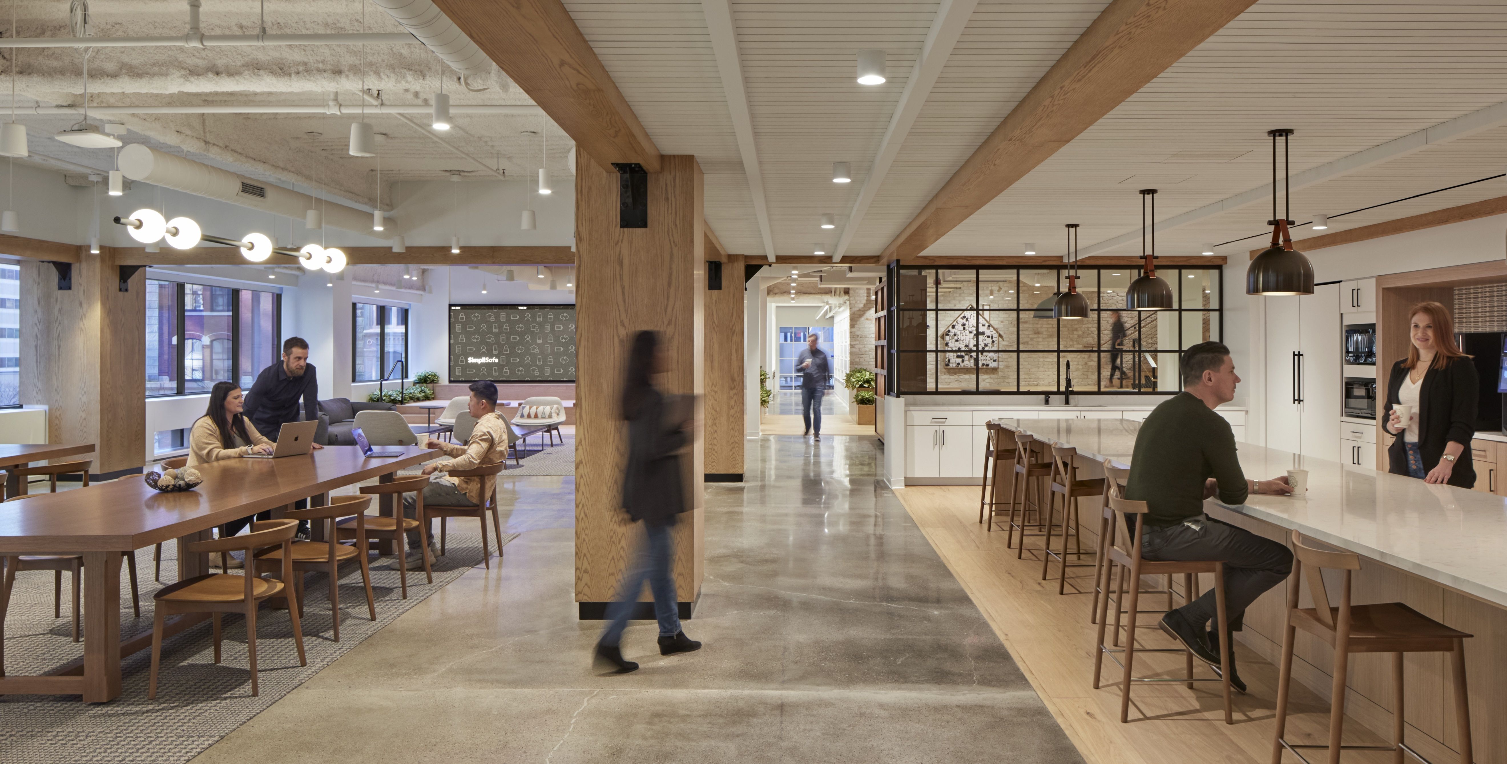 Employees are seen sitting at a long wooden table on the left of the dining area and a sleek white table on the right at SimpliSafe's HQ.