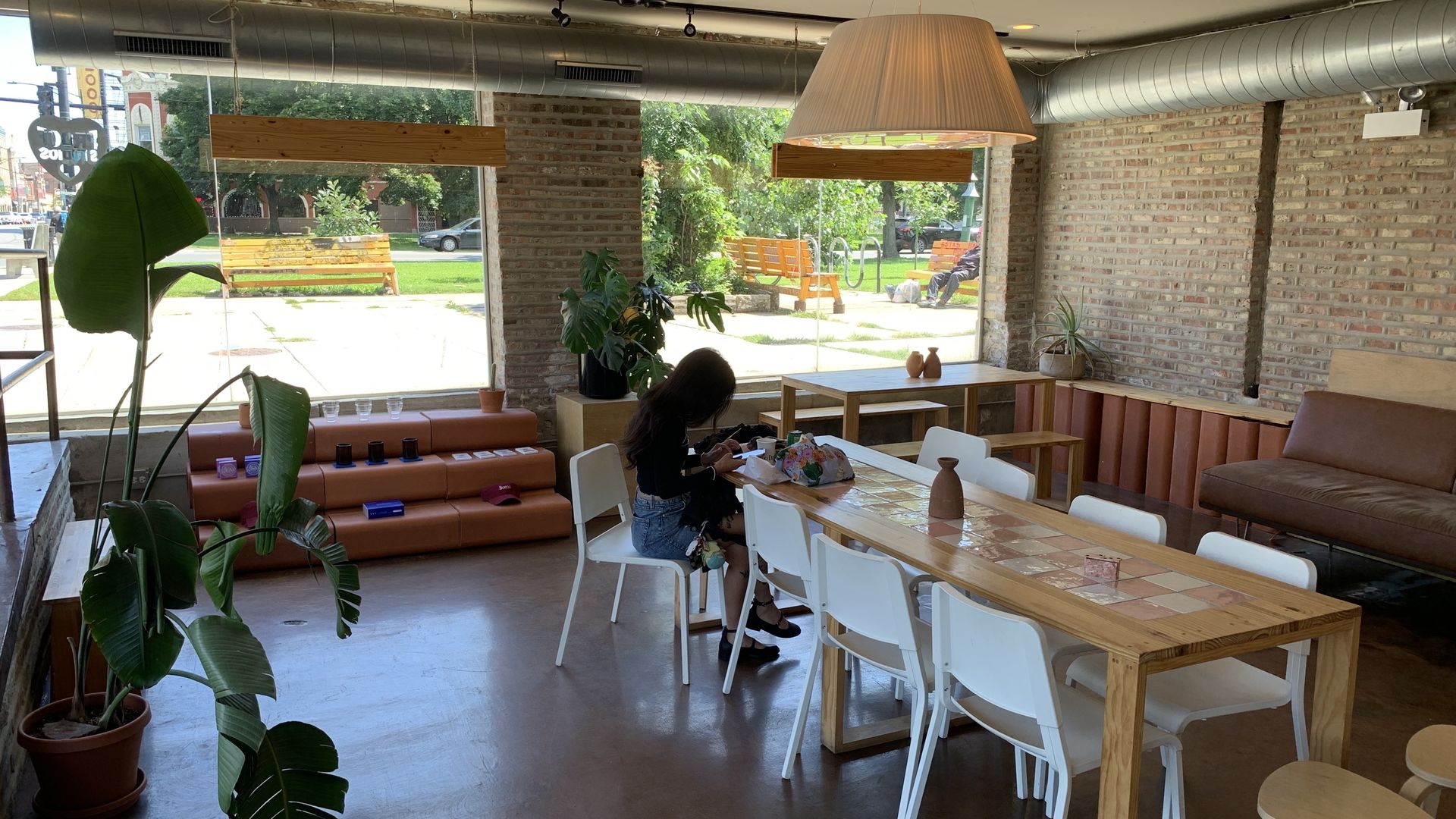 Cozy cafe interior with large windows showing green park outside, wooden table with white chairs, brown bench seating, large plant, and a person sitting and writing.