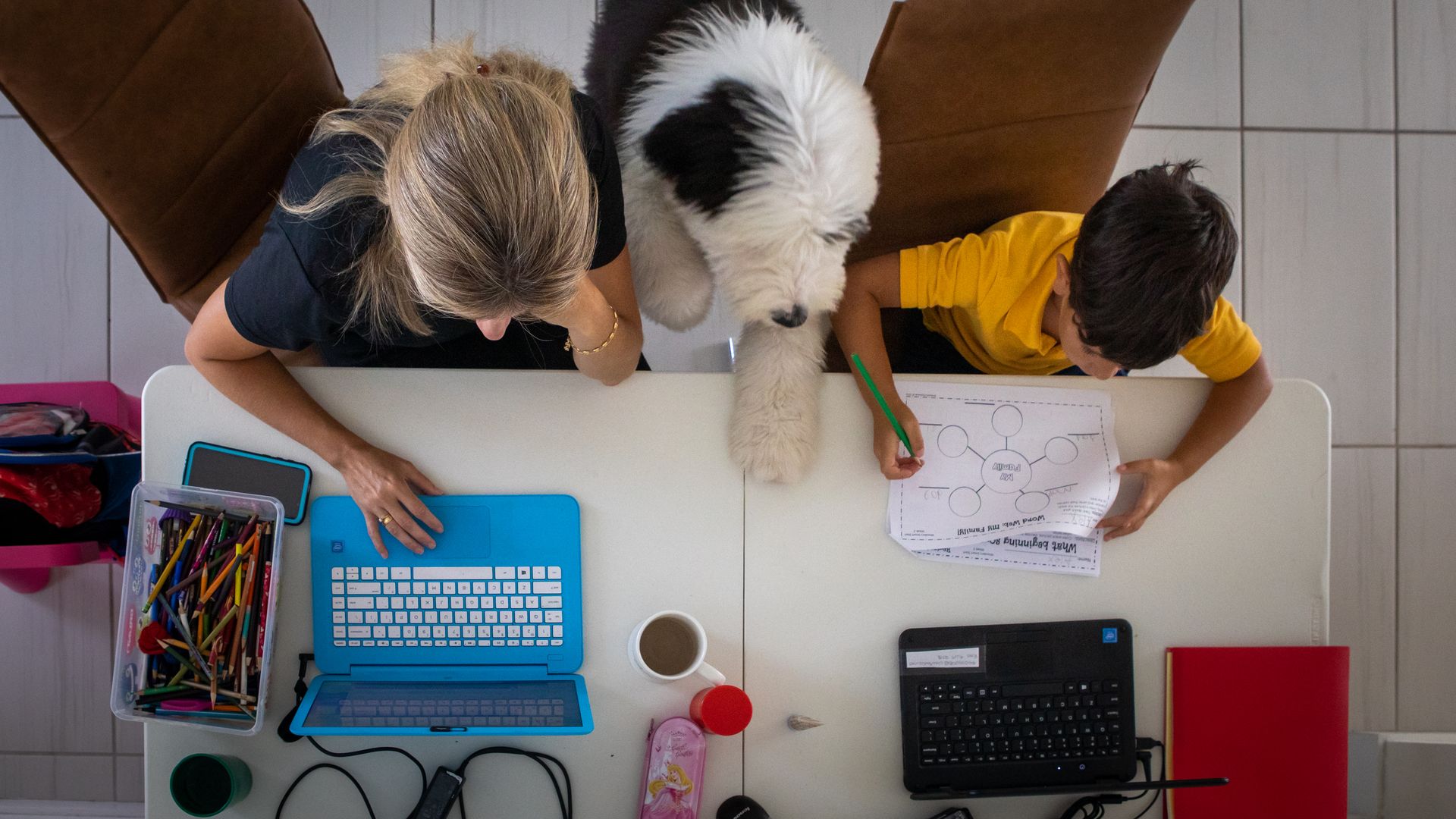 A woman and a boy work on various electronics at a table with a large dog between them.