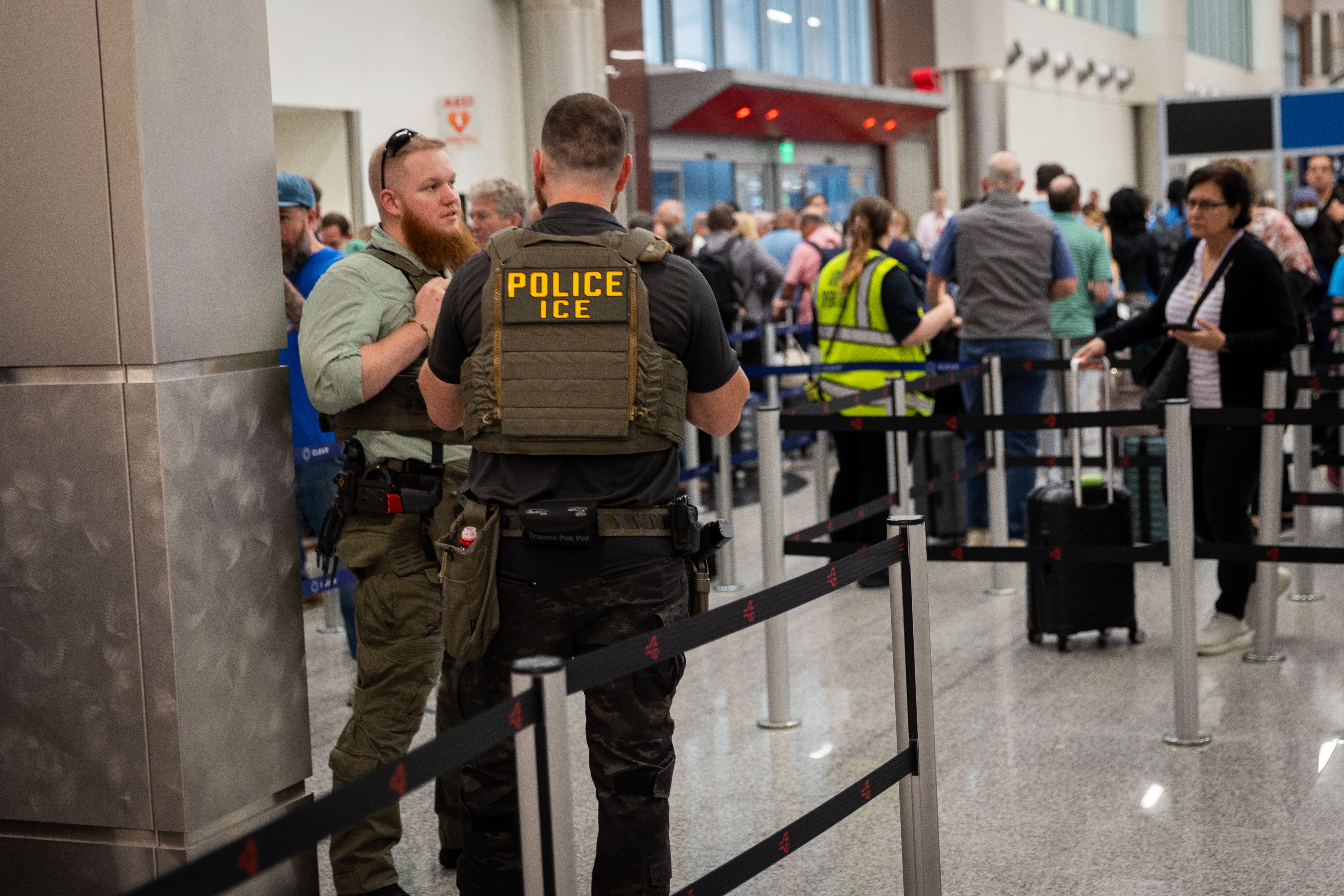 Two men wearing green tactical vests, one with the words "POLICE ICE" written on the back, talk as passengers wait in a security line at an airport.
