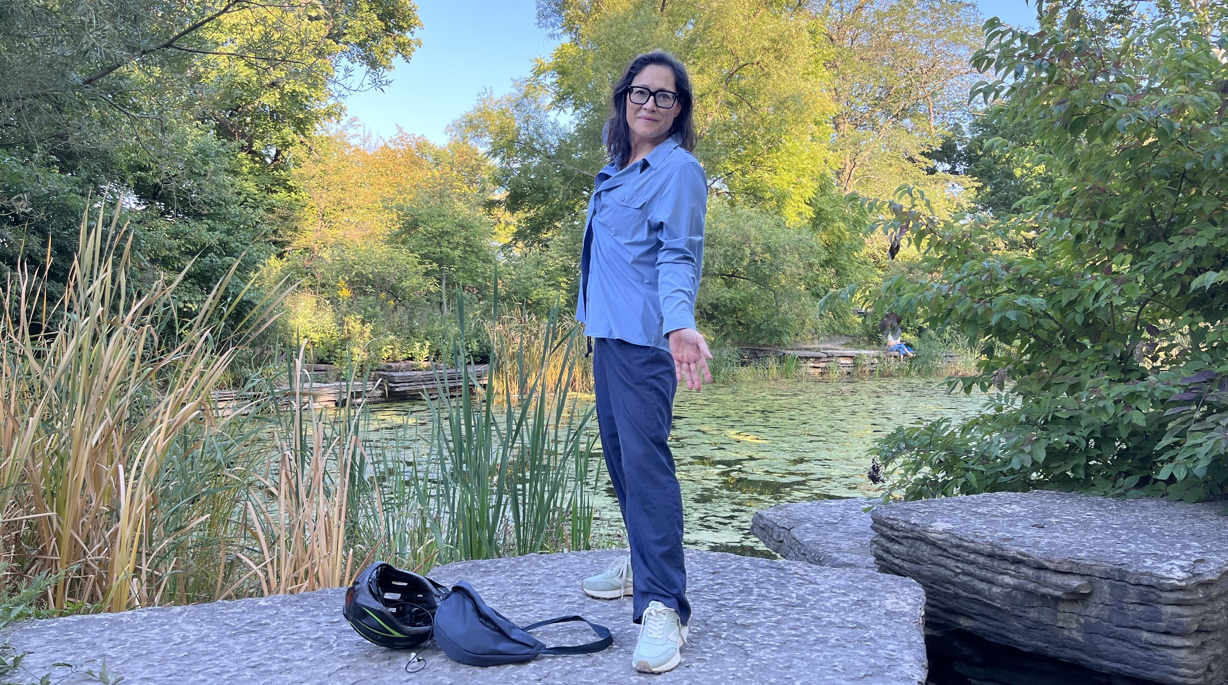 Woman in blue jacket and navy pants stands on large stone near a pond with green lily pads, surrounded by lush green and yellow trees under clear blue sky.