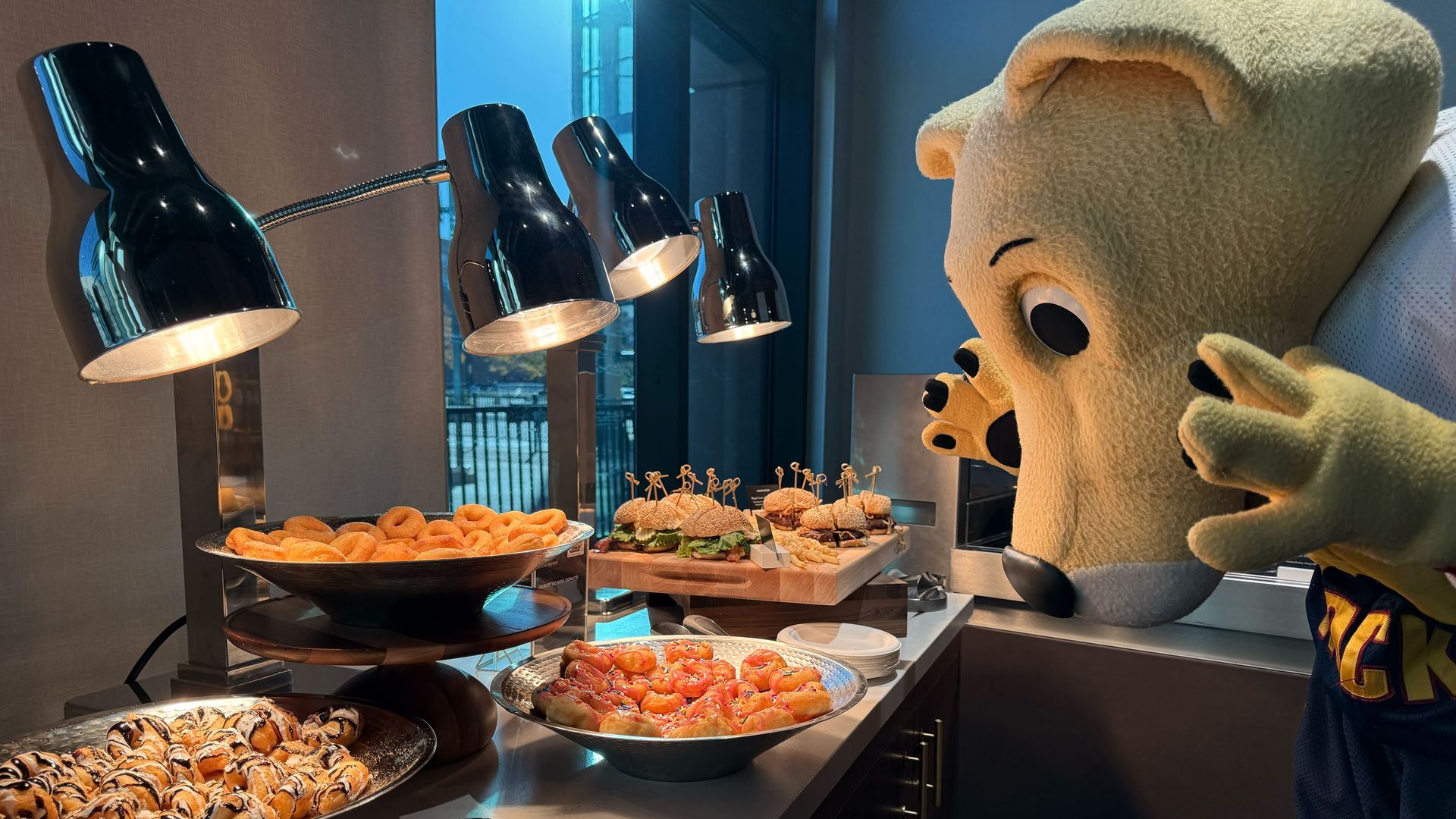 A yellow mascot character leans over a buffet table with food under lamps in a room with a windows. 