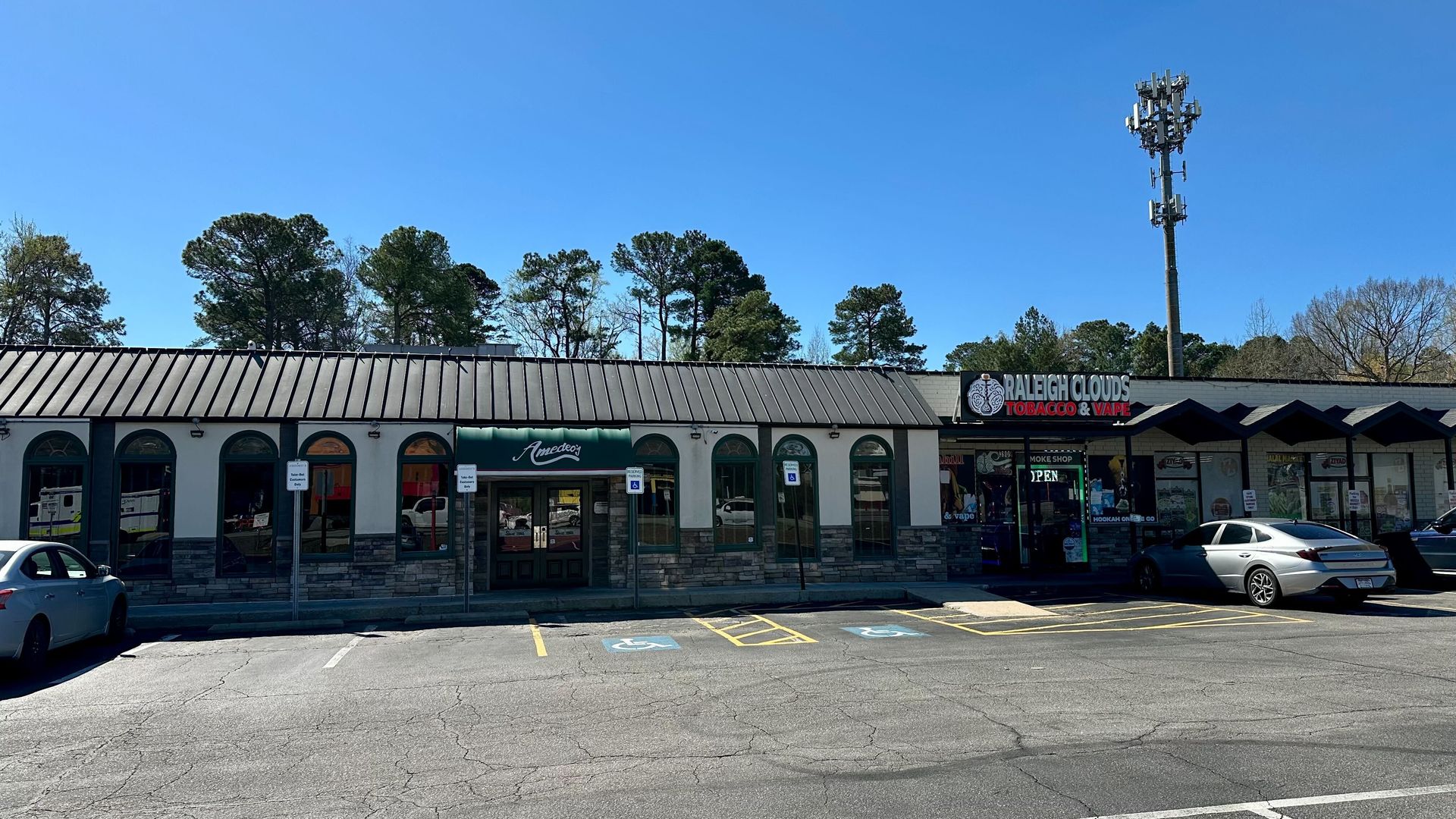 Single-story strip mall with arched windows and a stone base. Signs read Amedeo's and Raleigh Clouds Tobacco & Vape. Cracked parking lot with handicap spaces, a couple cars, a tall cell tower, blue sky, trees behind.