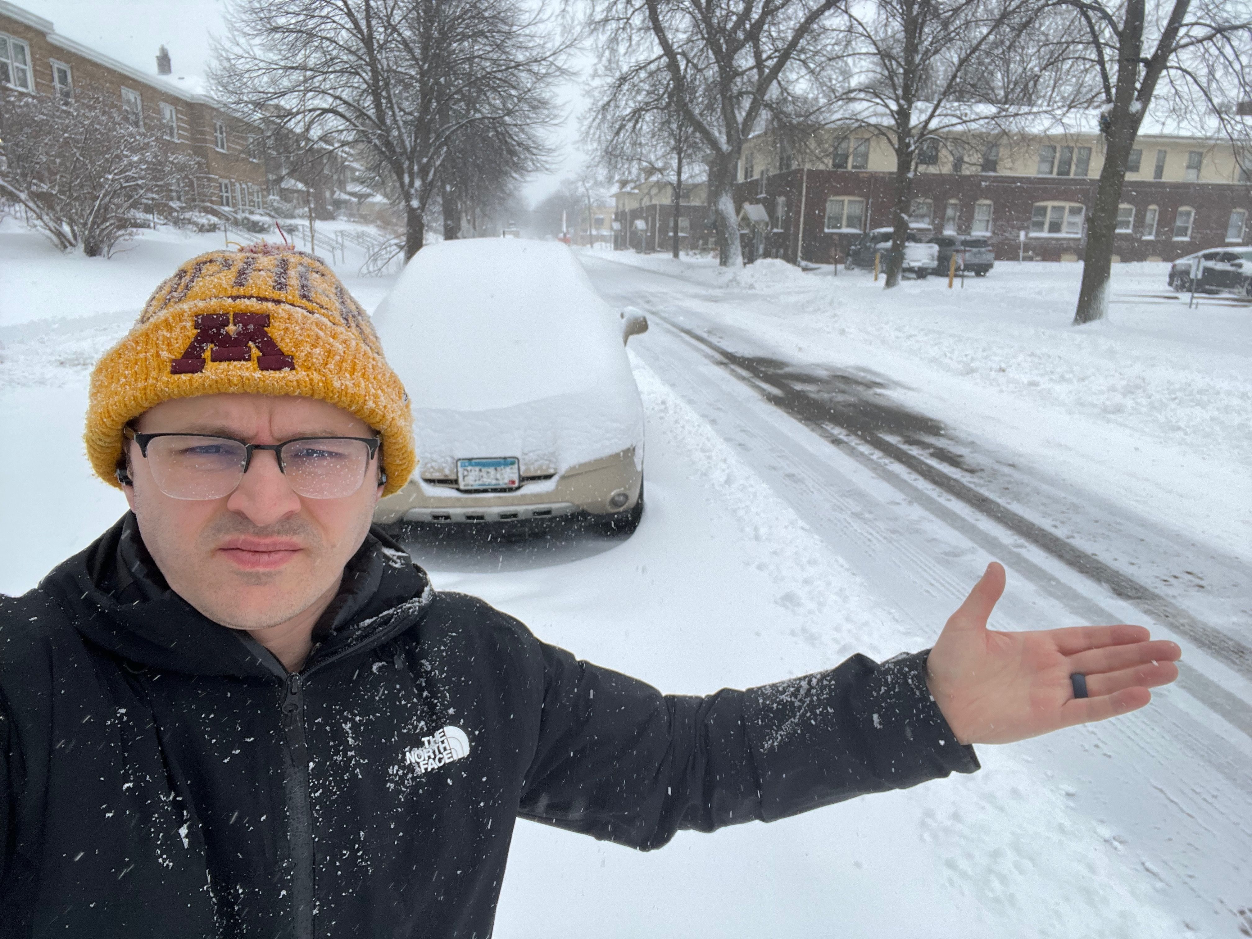 Self-portrait of a man in a black North Face jacket and a yellow knit hat with a maroon Minnesota logo, posing on a snowy residential street with a snow-covered car in the background.