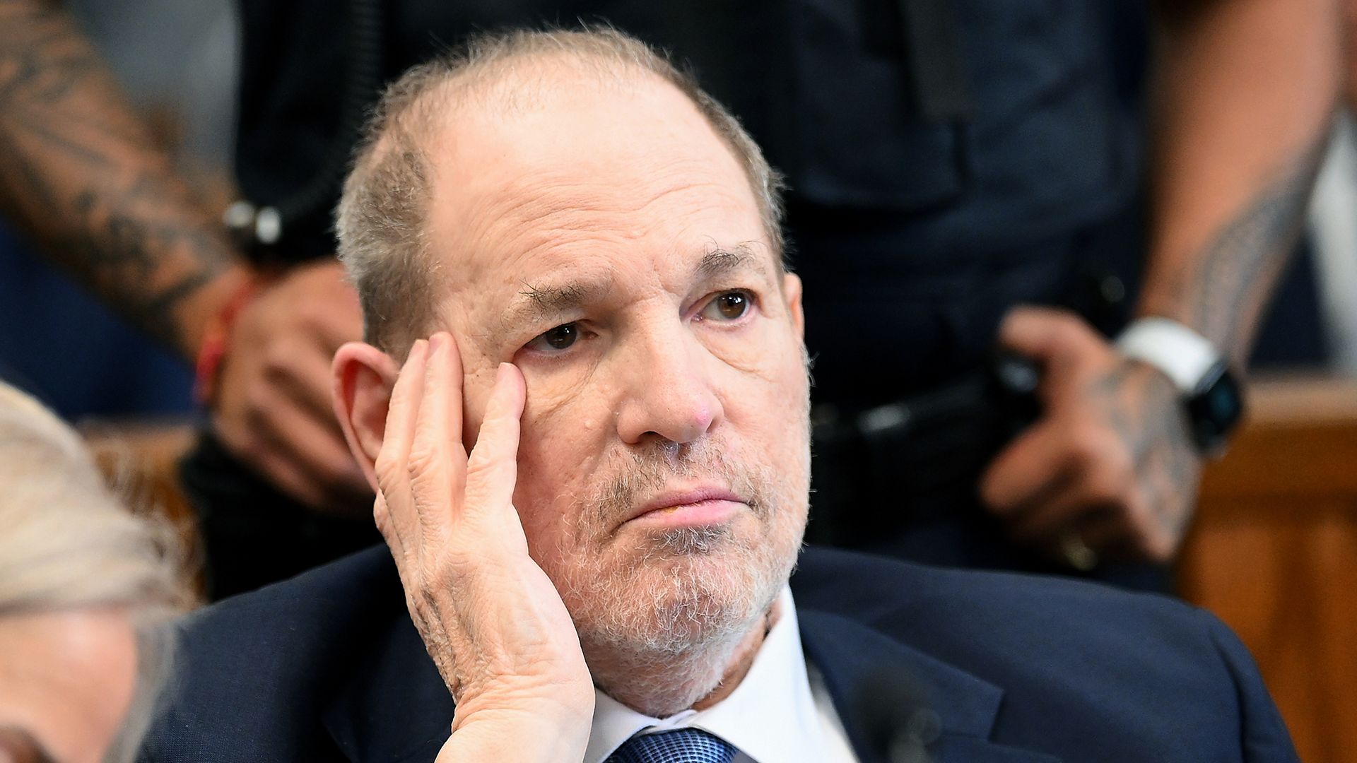 A close-up of Harvey Weinstein's face as he holds one cheek with his hand. He's wearing a dark suit and white shirt.