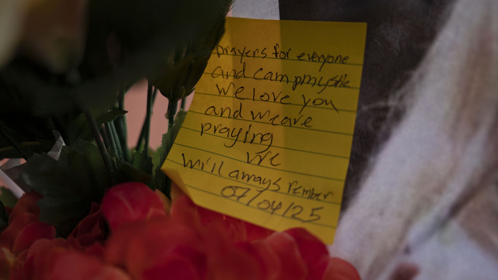 Close-up of a yellow sticky note with handwritten message about prayers and love, tucked among dark green stems and red flowers; a bouquet tribute.