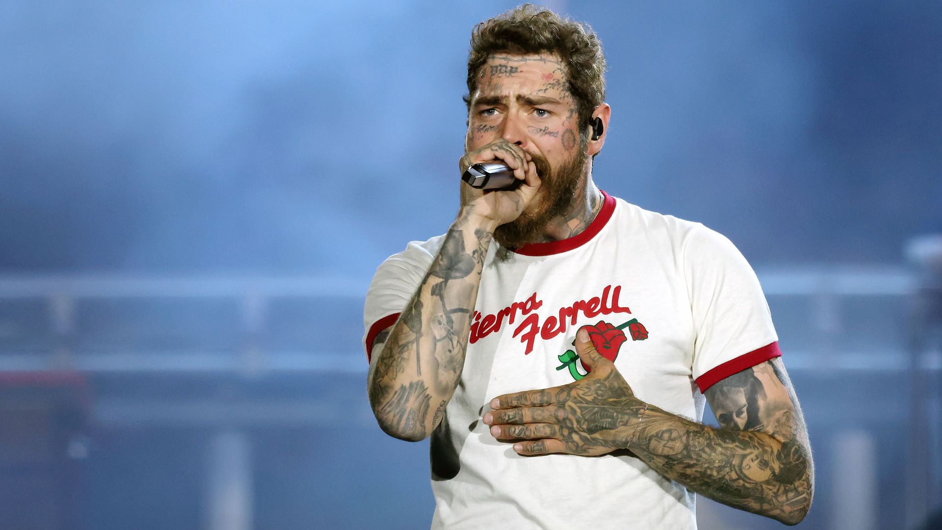 Man with tattoos on face and arms, wearing a white tee with red sleeves and "Sierra Ferrell" text, singing into a microphone on stage with a smoky blue background.