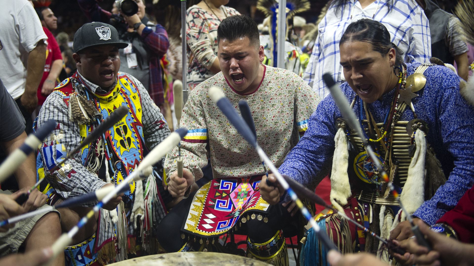 Three Indigenous men use large sticks to beat a drum in front of them, while they appear to sing while surrounded by other people. They are wearing colorful sweaters and shirts.