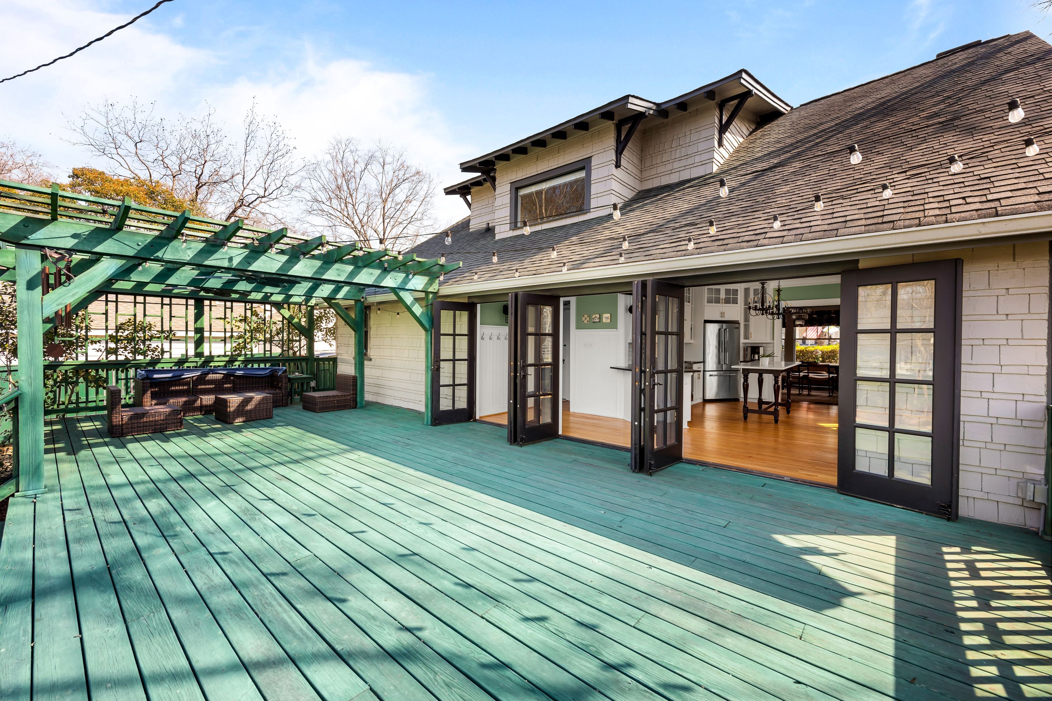 Spacious outdoor deck with teal wooden floor and green pergola with string lights, dark wood patio furniture, and open black-framed French doors leading to a light-filled kitchen and dining area.