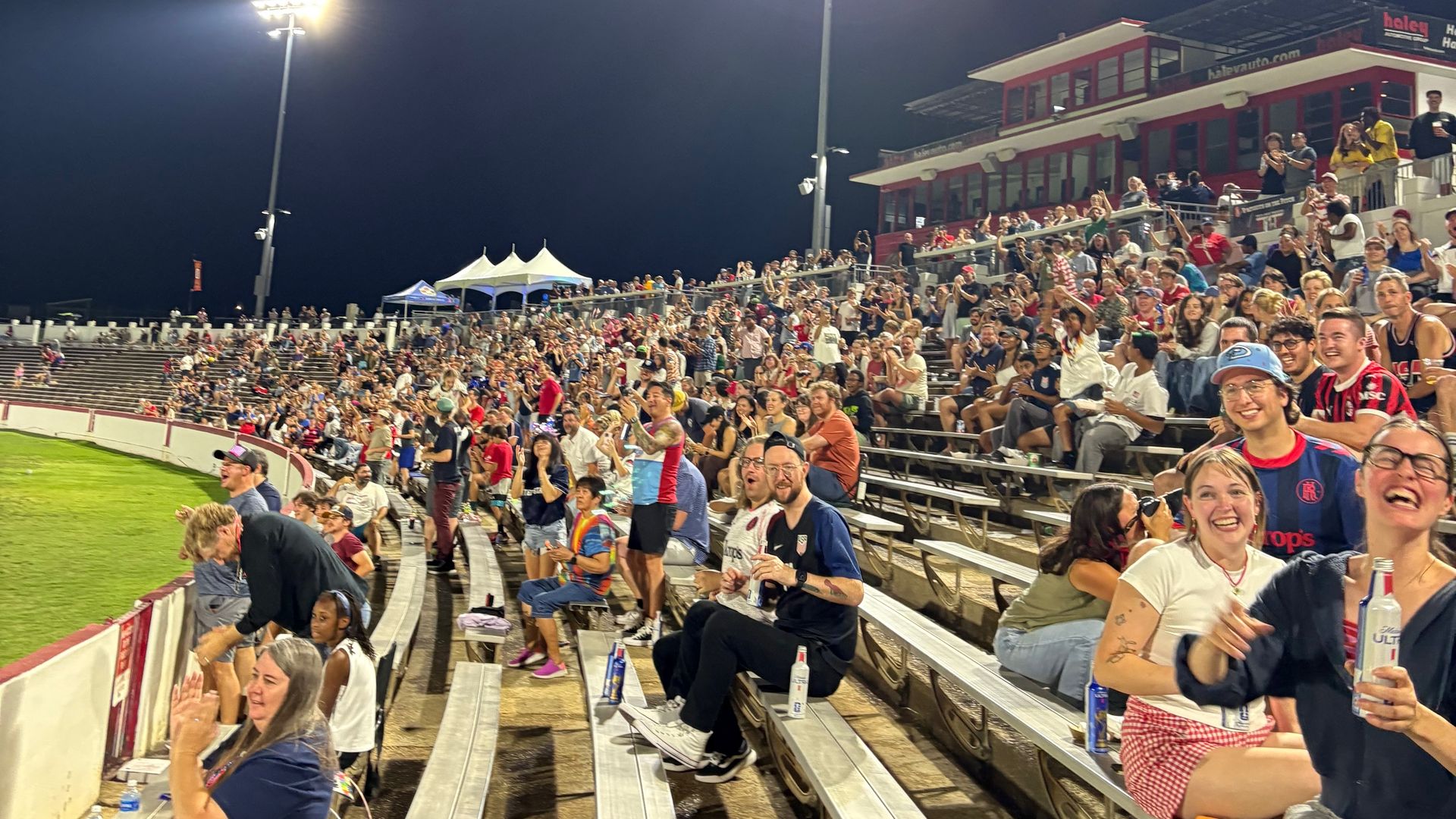 Nighttime stadium with crowded metal bleachers; bright floodlights on tall poles illuminate fans, a red multi-level press box to the right, and tents to the left beneath a dark sky.