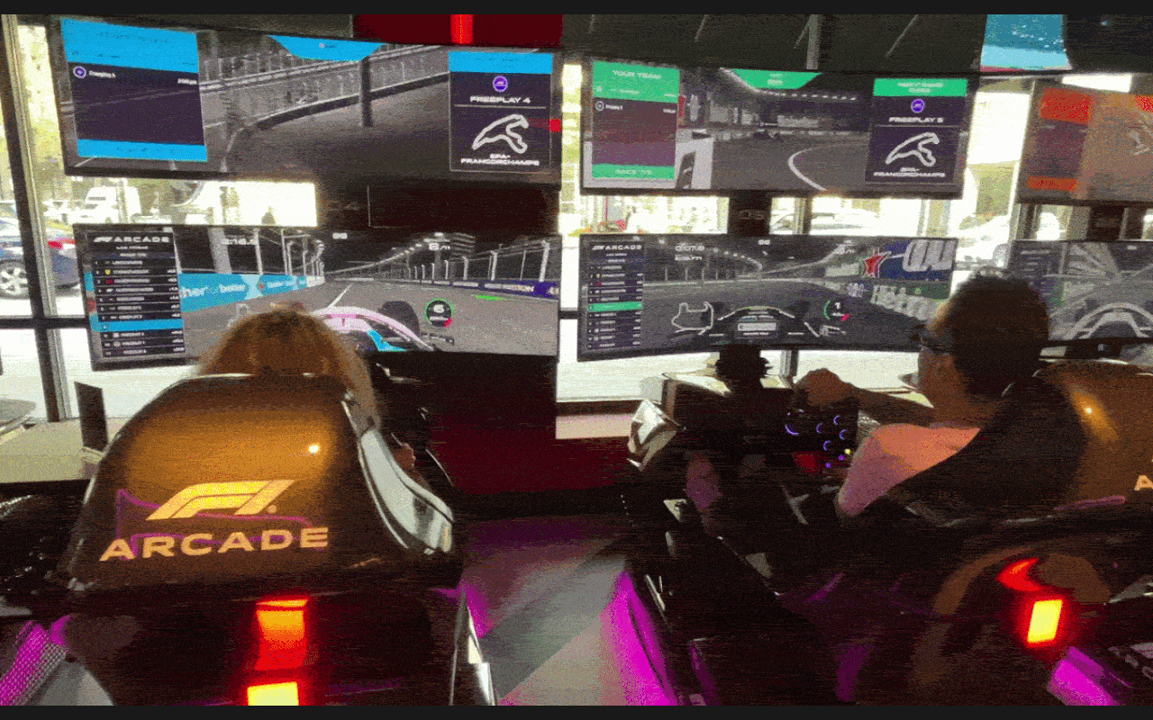Gif of two people sitting inside racecar simulators steering a wheel with screens showing a virtual racetrack