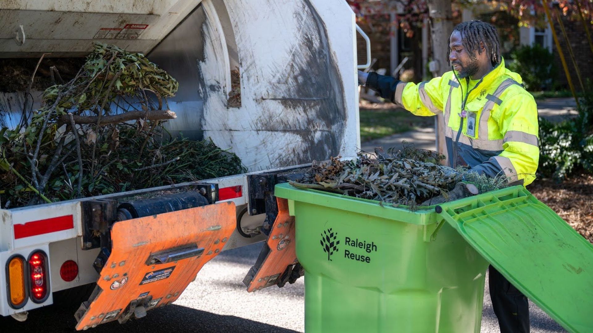 Worker in bright yellow jacket loading green Raleigh Reuse bin filled with branches and leaves into a white truck with orange mechanical loader on a sunny day.