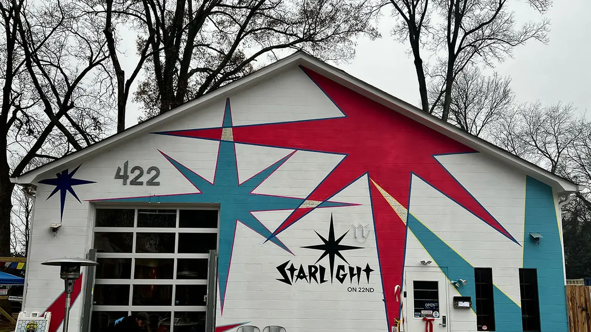 White building with large red and blue starburst mural, gray number 422, black Starlight logo, and glass garage door, set against leafless trees under overcast sky.