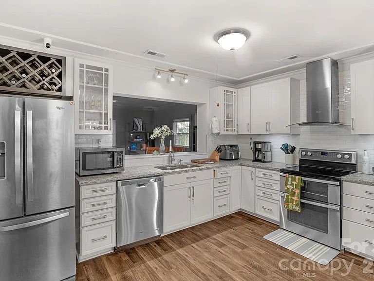 Modern kitchen with white cabinets, stainless steel refrigerator, dishwasher, stove, microwave, hardwood floor, and granite countertops; kitchen opens to a living room through a pass-through window.