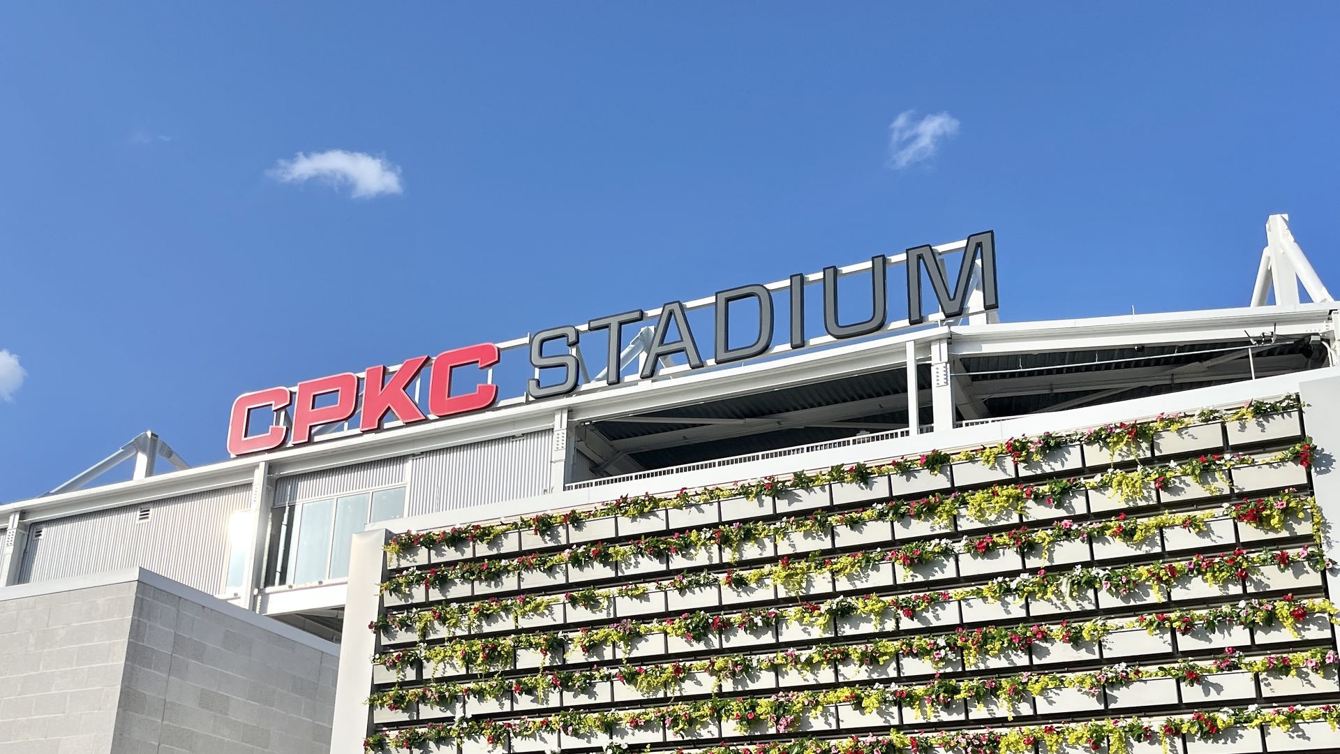 Blue sky above a modern stadium building with a red CPKC sign and dark STADIUM letters, and rows of flower-filled planters on the facade.