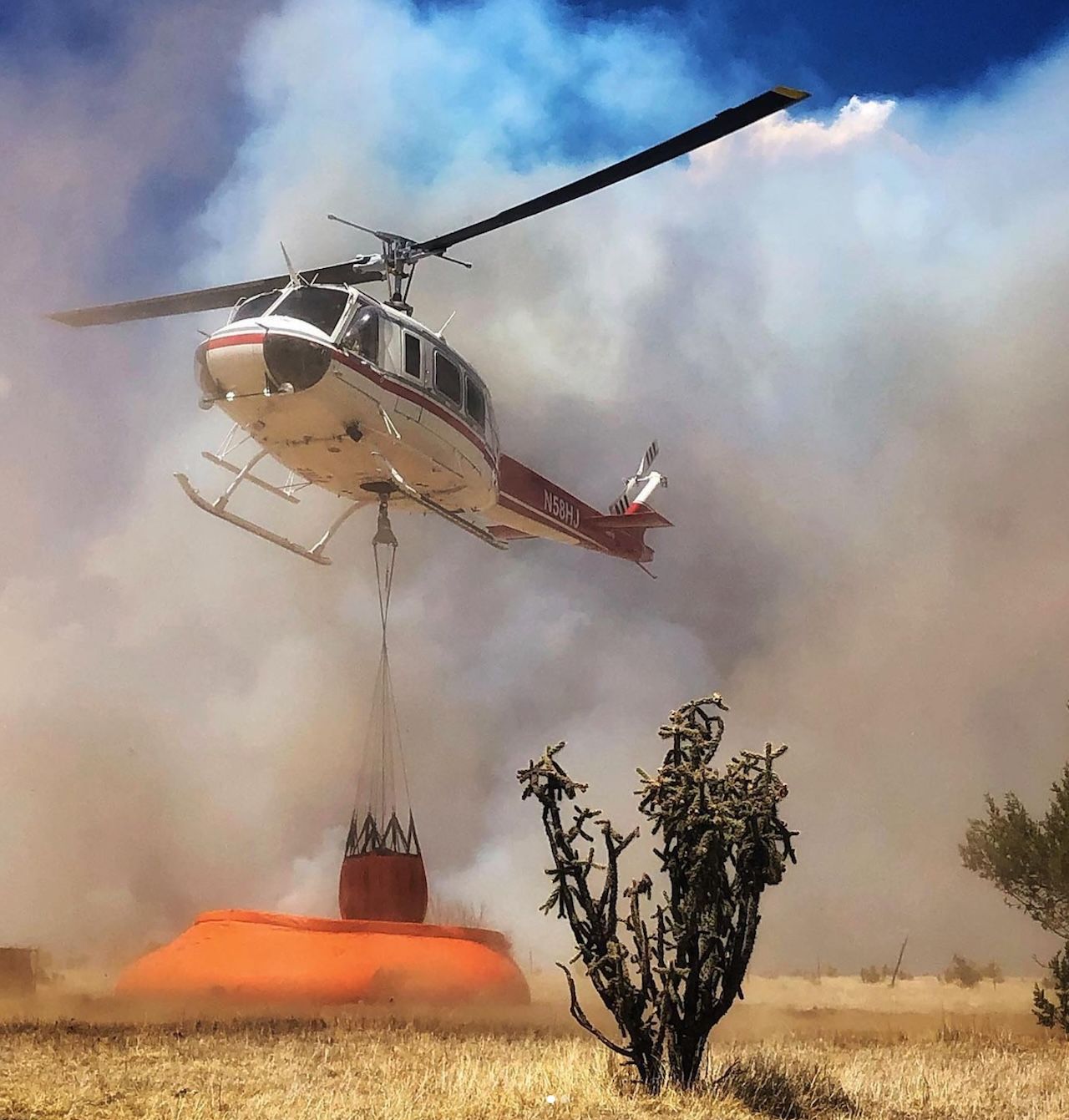 A white and red firefighting helicopter hovers in a smoky sky, lowering a large orange water bucket toward a dry field; a long cable suspends the bucket beside sparse scrub in the foreground.