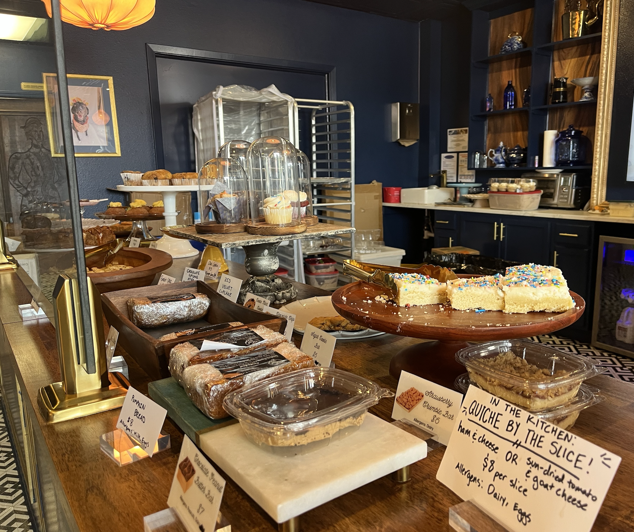 Sweet bakery counter with cupcakes under glass domes, assorted pastries on a wooden table; blue walls, gold-framed art, orange pendant lamp, and shelves with blue glass bottles in the background.
