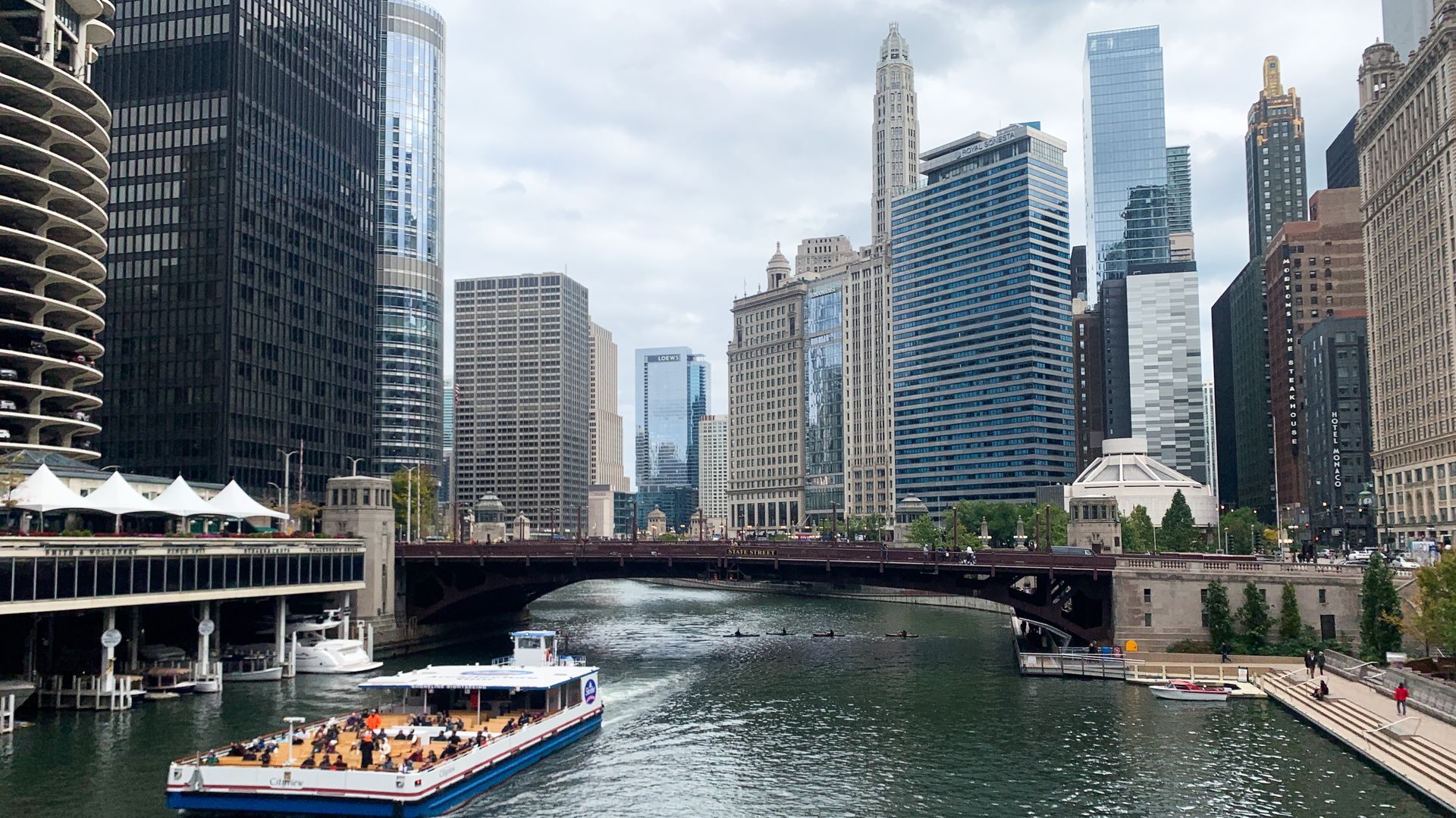 Tour boat on Chicago River with downtown Chicago skyscrapers and State Street bridge under cloudy sky, people on riverwalk and boats on water