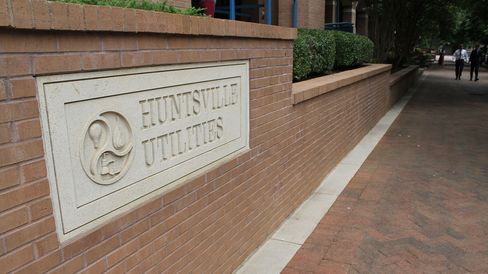 Stone sign reading "HUNTSVILLE UTILITIES" embedded in a brick wall next to a sidewalk with greenery and people walking in the background.