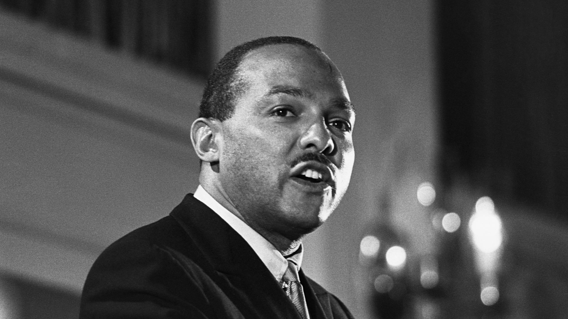 Black and white photo of a man in a suit speaking, captured from the side, with blurred lights and architectural background.