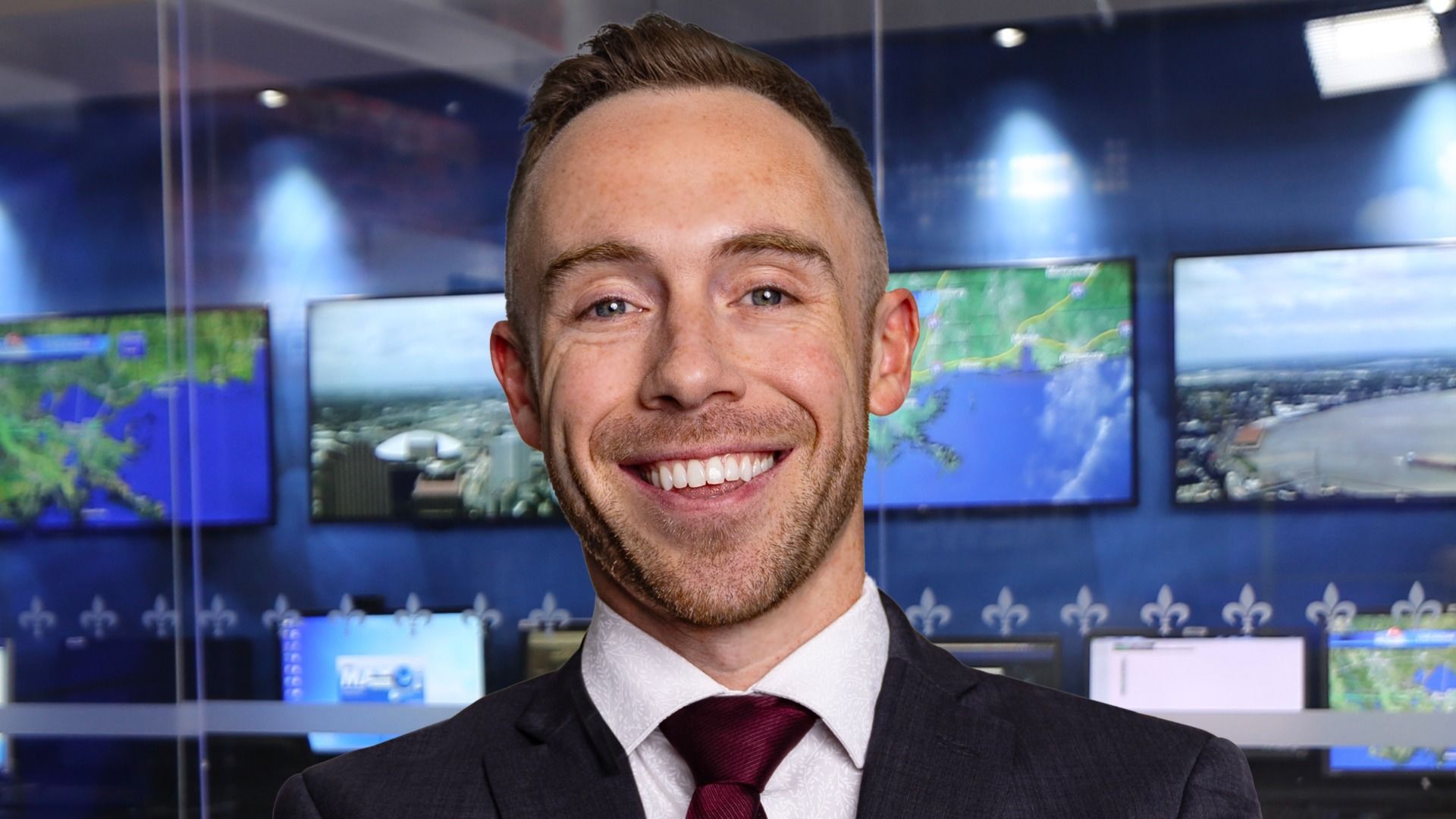 Smiling man in a dark suit and red tie poses in a newsroom with blue walls and multiple screens showing maps and cityscapes behind him.