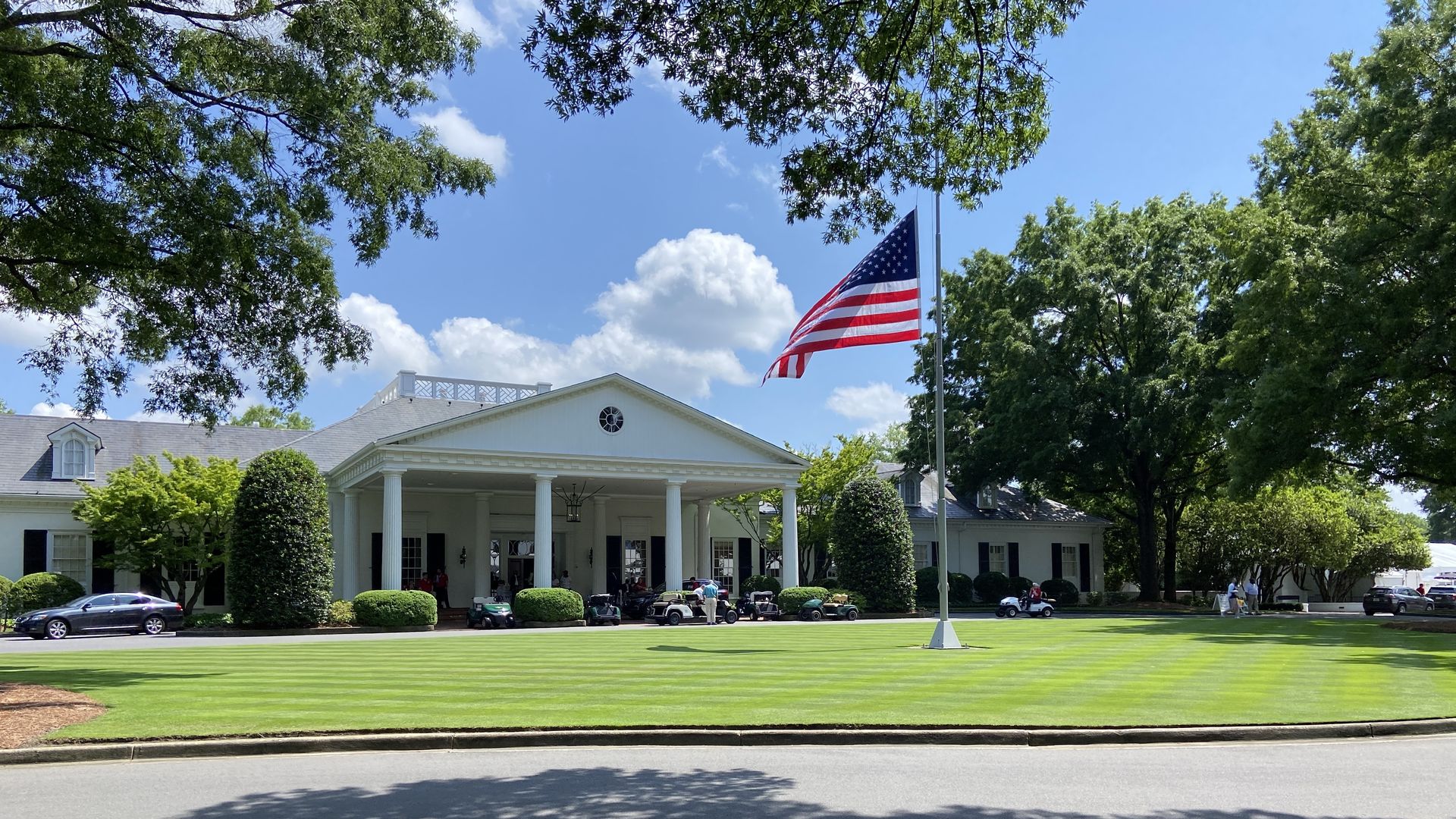 White building with columns, green lawn with striped mowing pattern, American flag at half-mast, golf carts, and trees on a sunny day with blue sky and clouds.