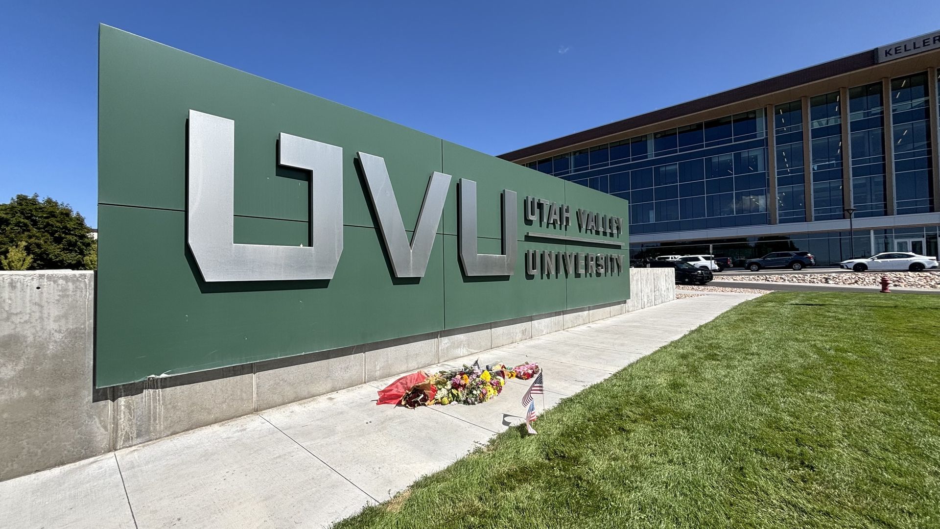 Large green sign with silver letters reading "UVU Utah Valley University" beside a sidewalk with flower bouquets and small American flags, near a modern glass building under a clear blue sky.
