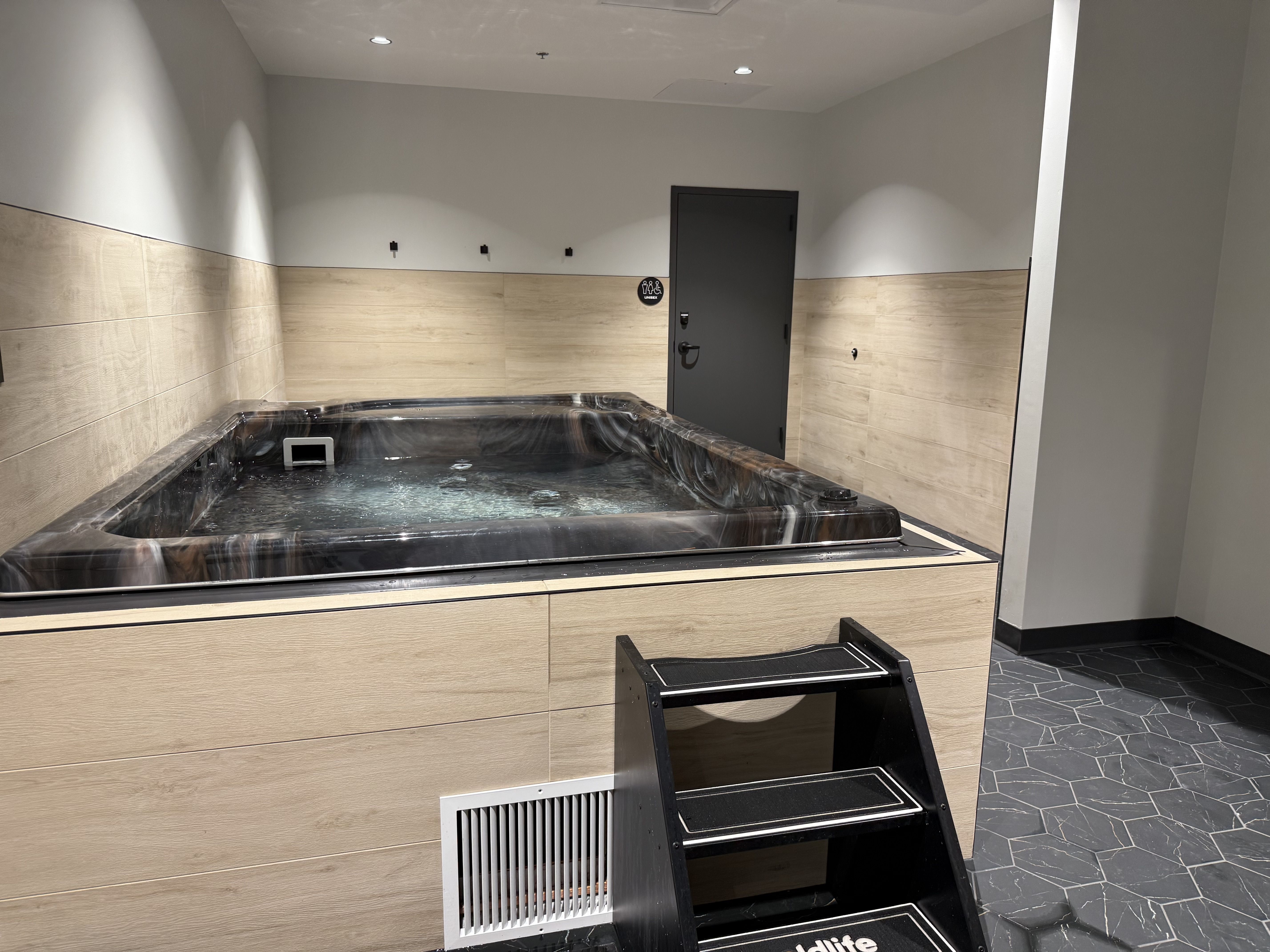 Indoor spa room featuring a dark marble hot tub against beige wood-tile walls, gray upper walls, and a dark door. Three wall hooks, a handicap sign, and a black step ladder in front of the tub.