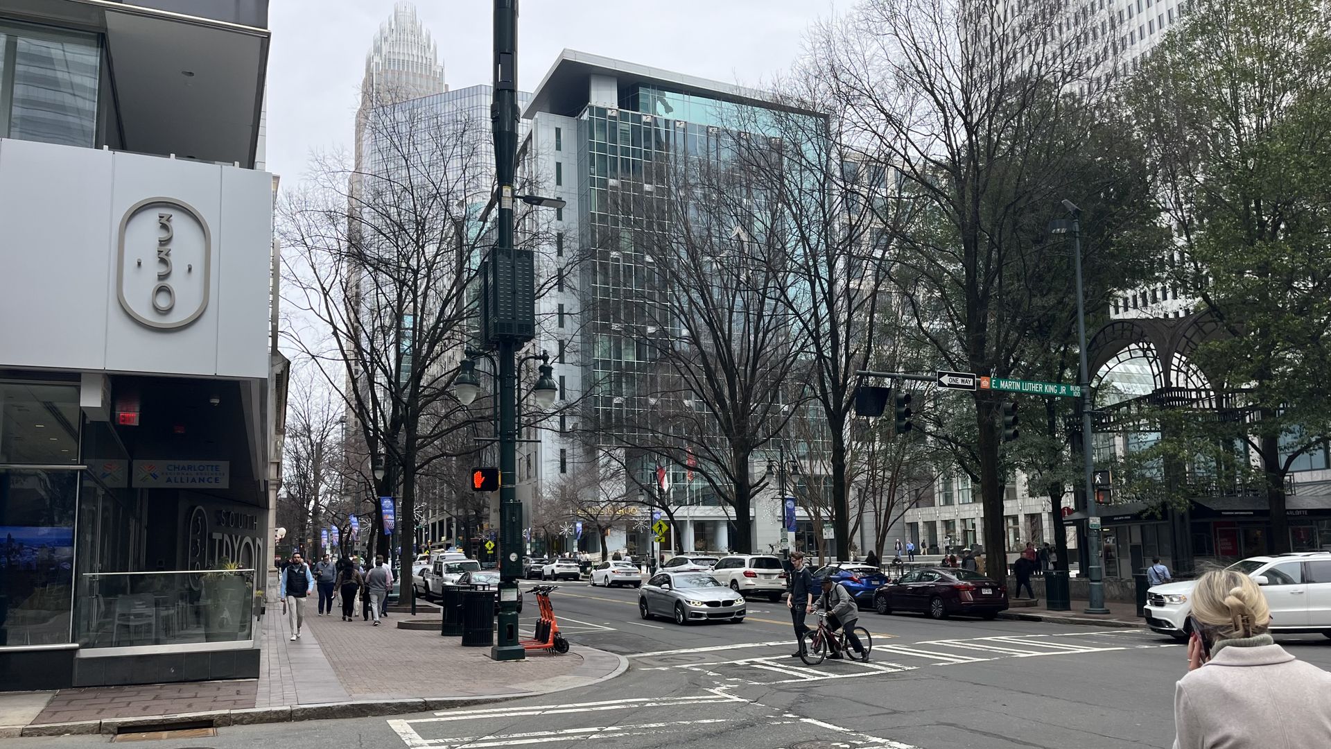 City street corner with tall glass buildings, leafless trees, and a crosswalk. A beige-coated woman on the right talks on her phone; a cyclist with a child crosses as cars line the street.
