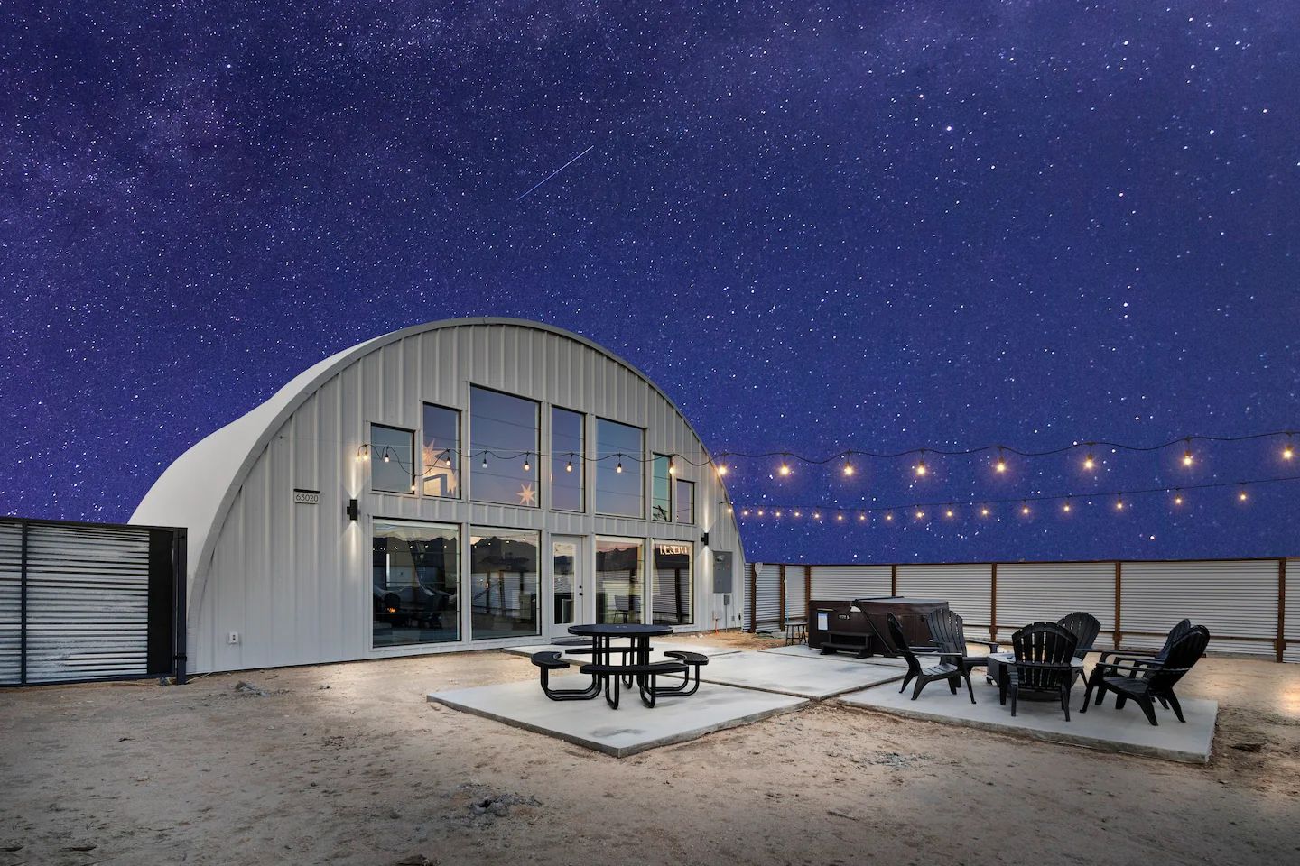 An airplane hangar converted into an airbnb has patio furniture, a hot tub and a fire pit in its fenced yard in the desert under a starry night sky.