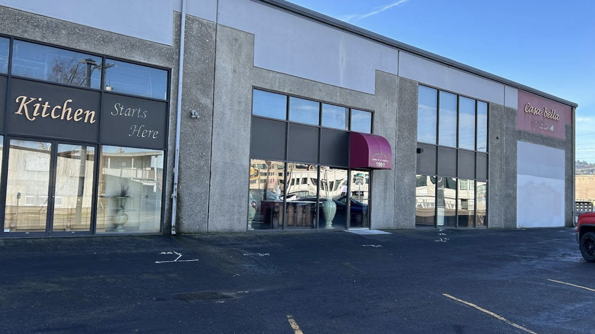 A commercial building with large glass windows houses "Casa Bella" and "Kitchen Starts Here" businesses. A red awning marks the entrance at 1901, and a red pickup truck is parked outside under clear skies. 