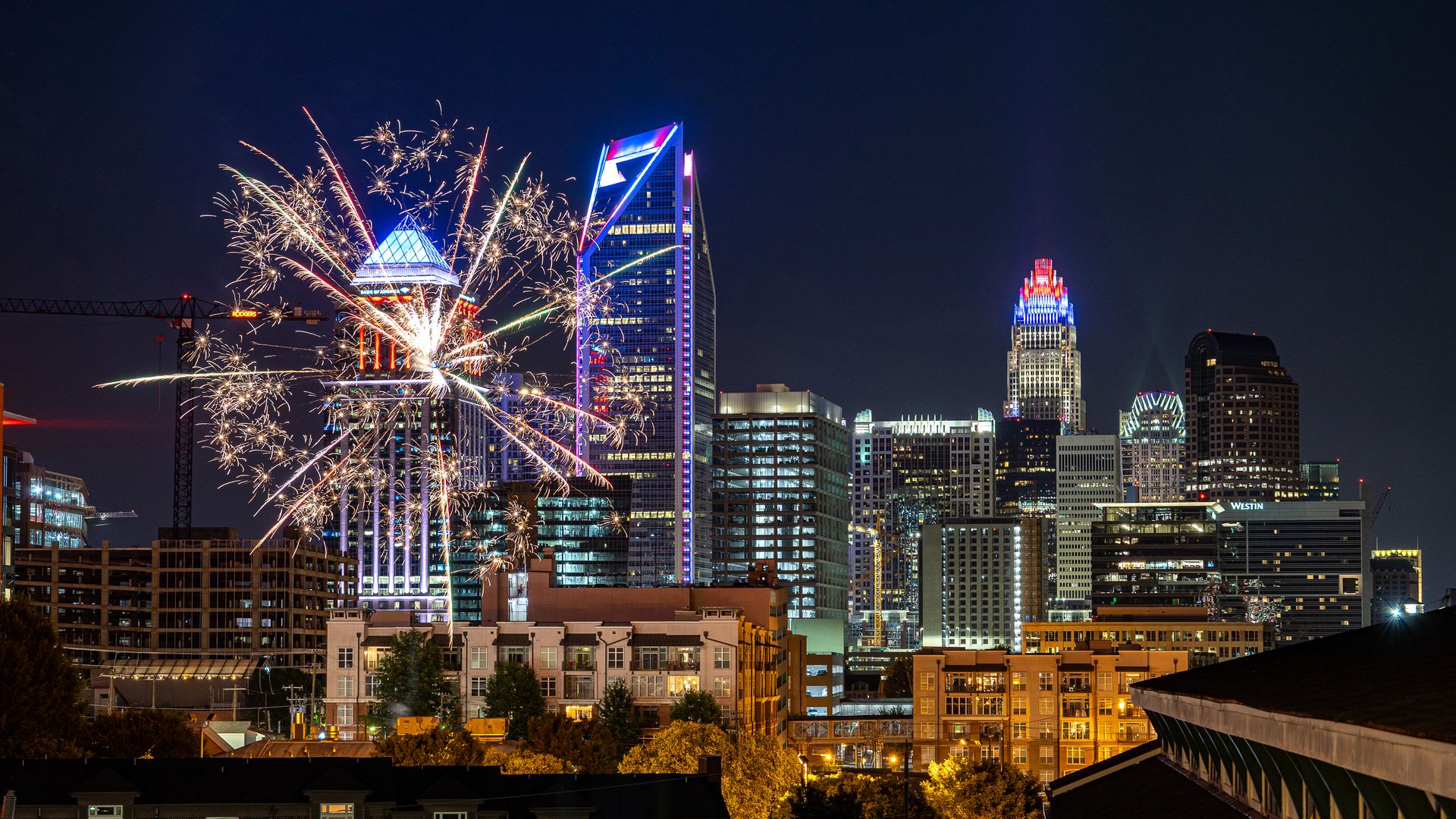 Nighttime city skyline with colorful fireworks bursting above tall, illuminated skyscrapers; a glass pyramid-roofed building and blue neon accents anchor the center.