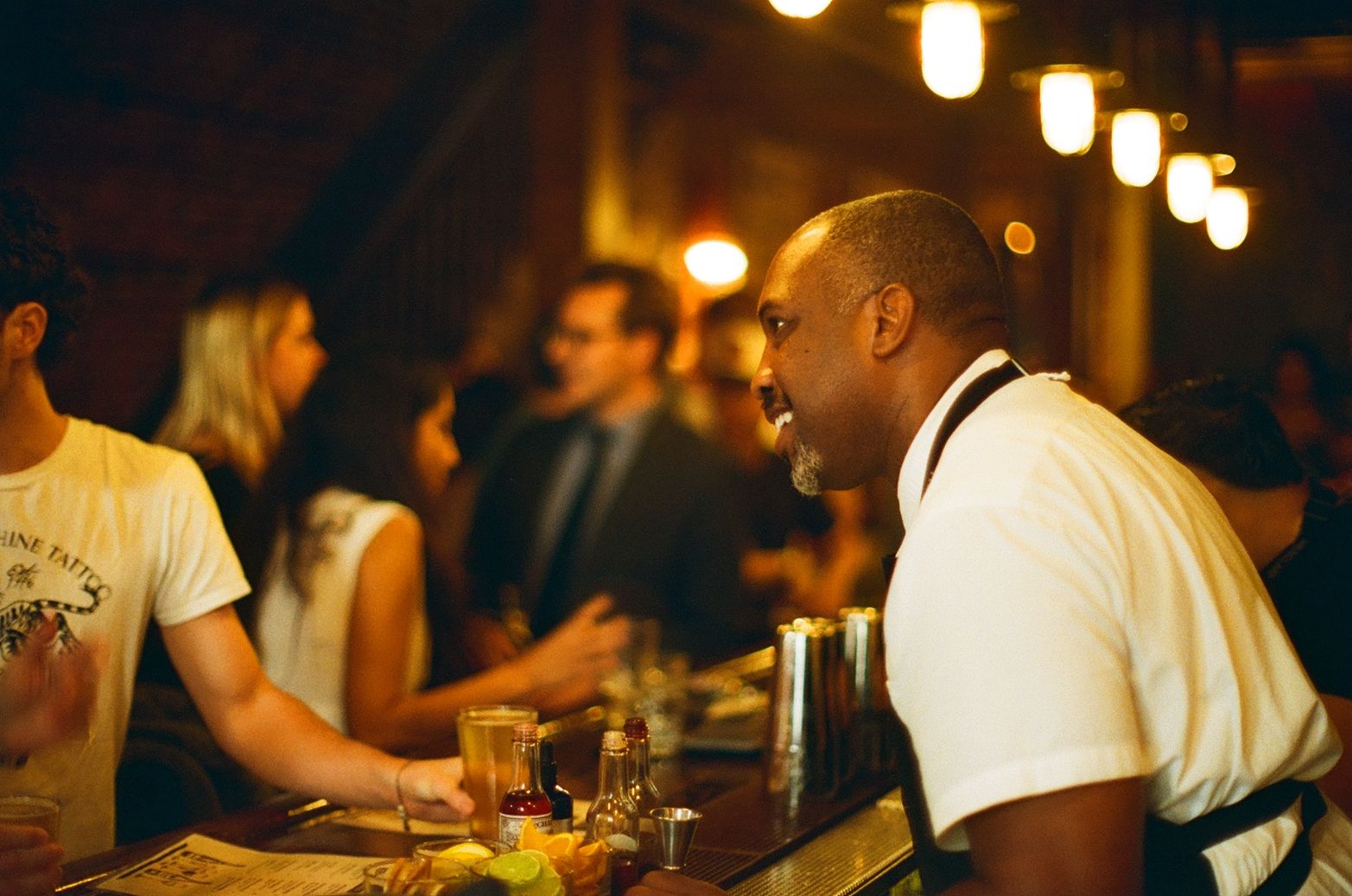 Chef Rock Harper in white shirt and black apron smiling and talking to a customer at a dimly lit bar with drinks and garnishes on the counter. Several other patrons and warm lights in the background.
